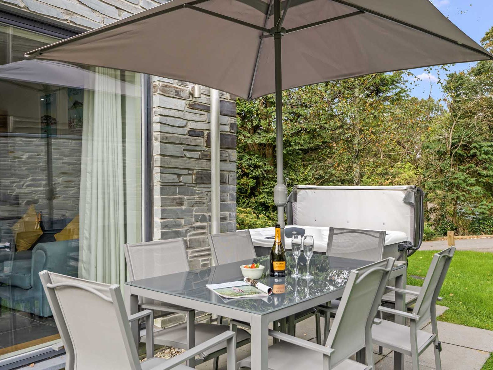 Outdoor patio with glass table, chairs, large umbrella, hot tub, and green surroundings.