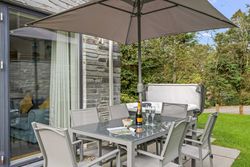 Outdoor patio with glass table, chairs, large umbrella, hot tub, and green surroundings.