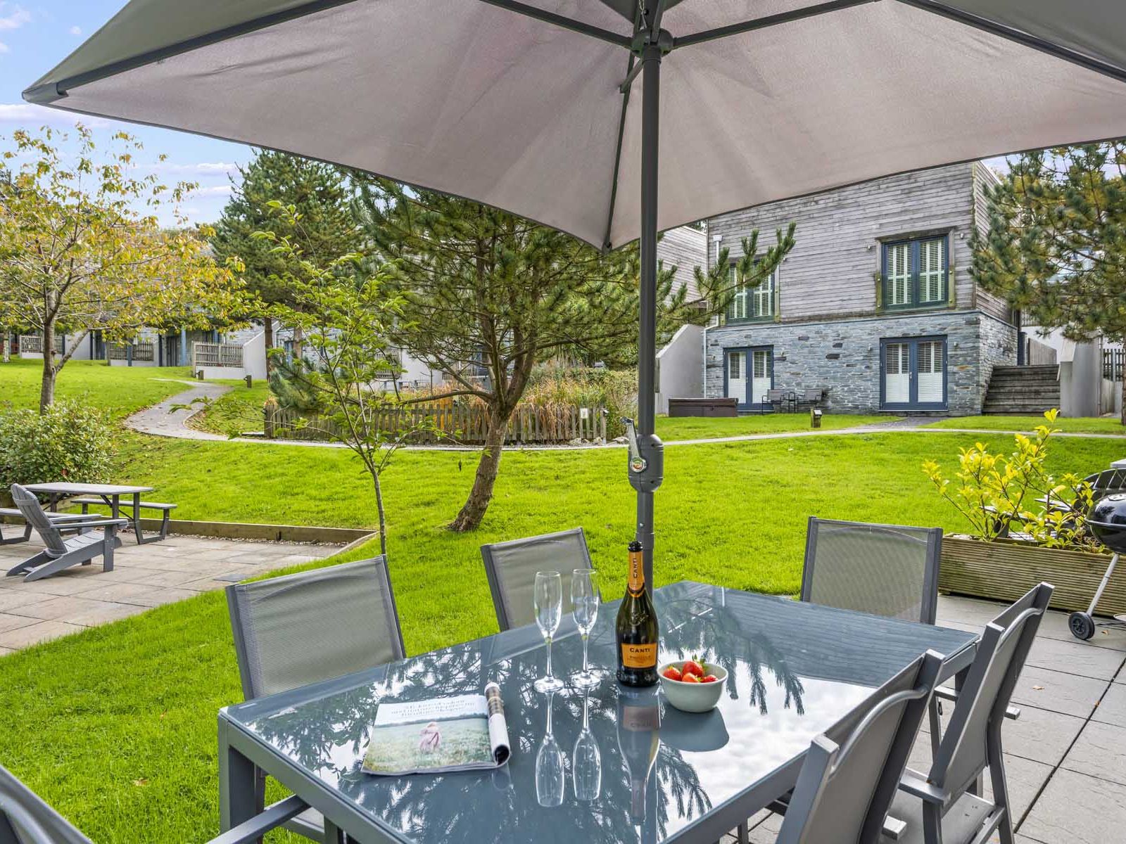 Outdoor patio with dining table, chairs, champagne, and bowl of strawberries under a large umbrella, overlooking a grassy yard with modern buildings.