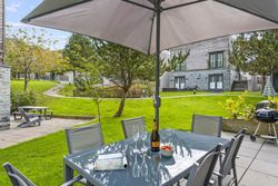 Outdoor patio with dining table, chairs, champagne, and bowl of strawberries under a large umbrella, overlooking a grassy yard with modern buildings.