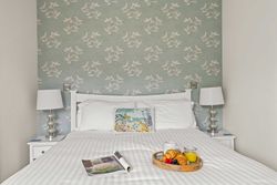 Breakfast tray with croissants, juice, and fruit on a neatly made bed with white linens, flanked by two lamps and a bird-patterned wallpaper.