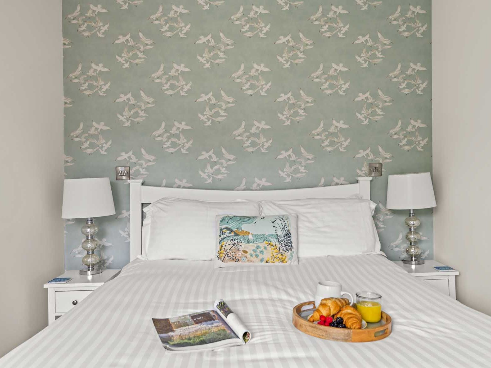 Breakfast tray with croissants, juice, and fruit on a neatly made bed with white linens, flanked by two lamps and a bird-patterned wallpaper.