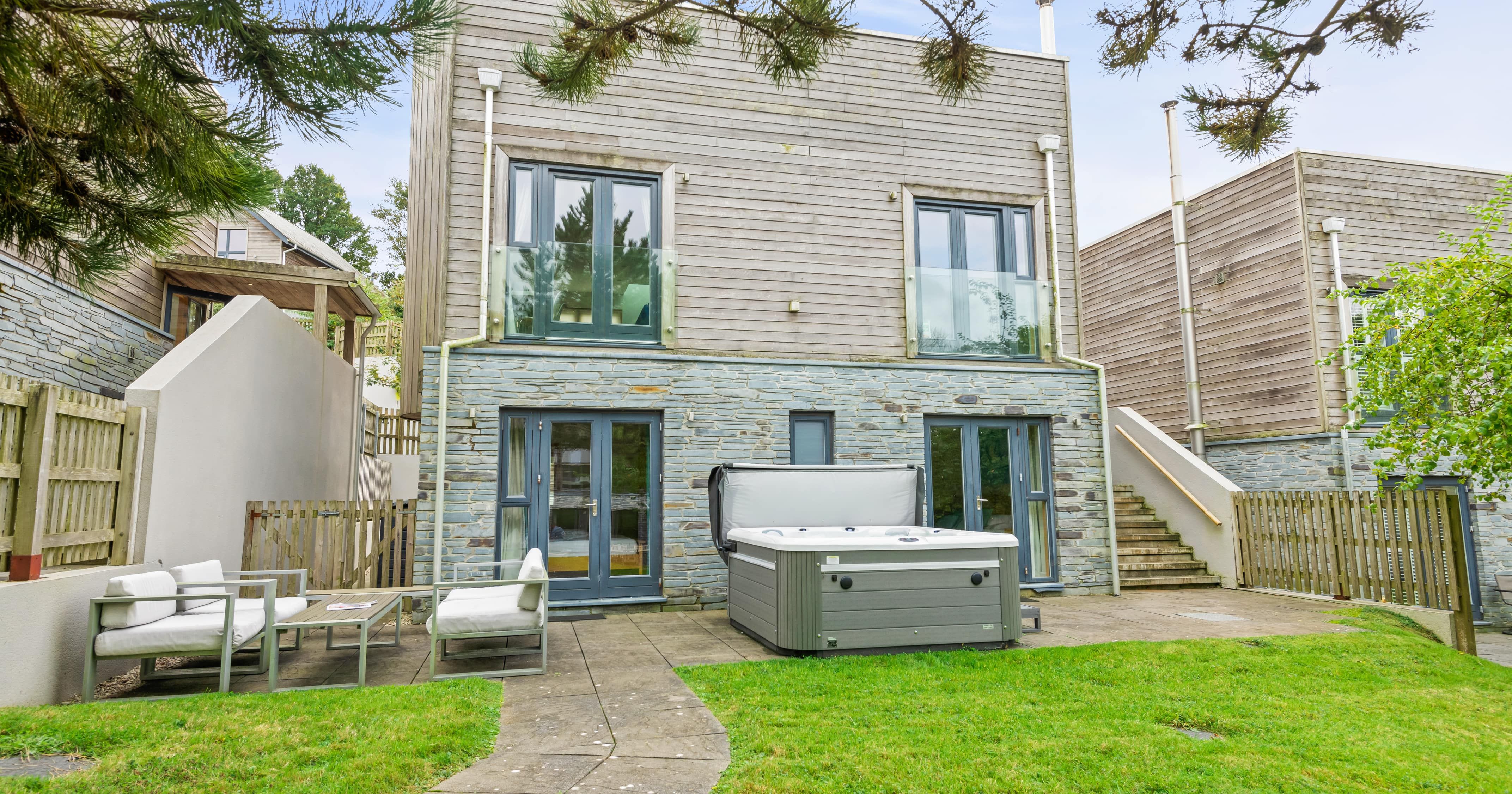 Modern two-story house with stone and wood facade, glass balconies, outdoor hot tub, patio furniture, green lawn, and surrounding wooden fencing.