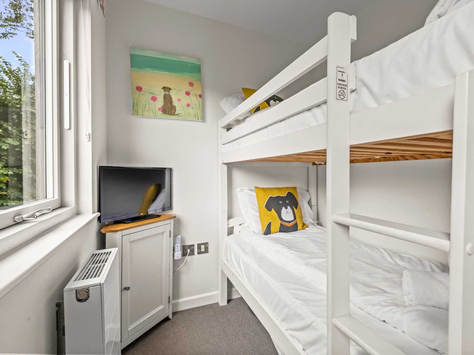 Small bedroom with white bunk beds, dog-themed pillow, wall art, and TV.