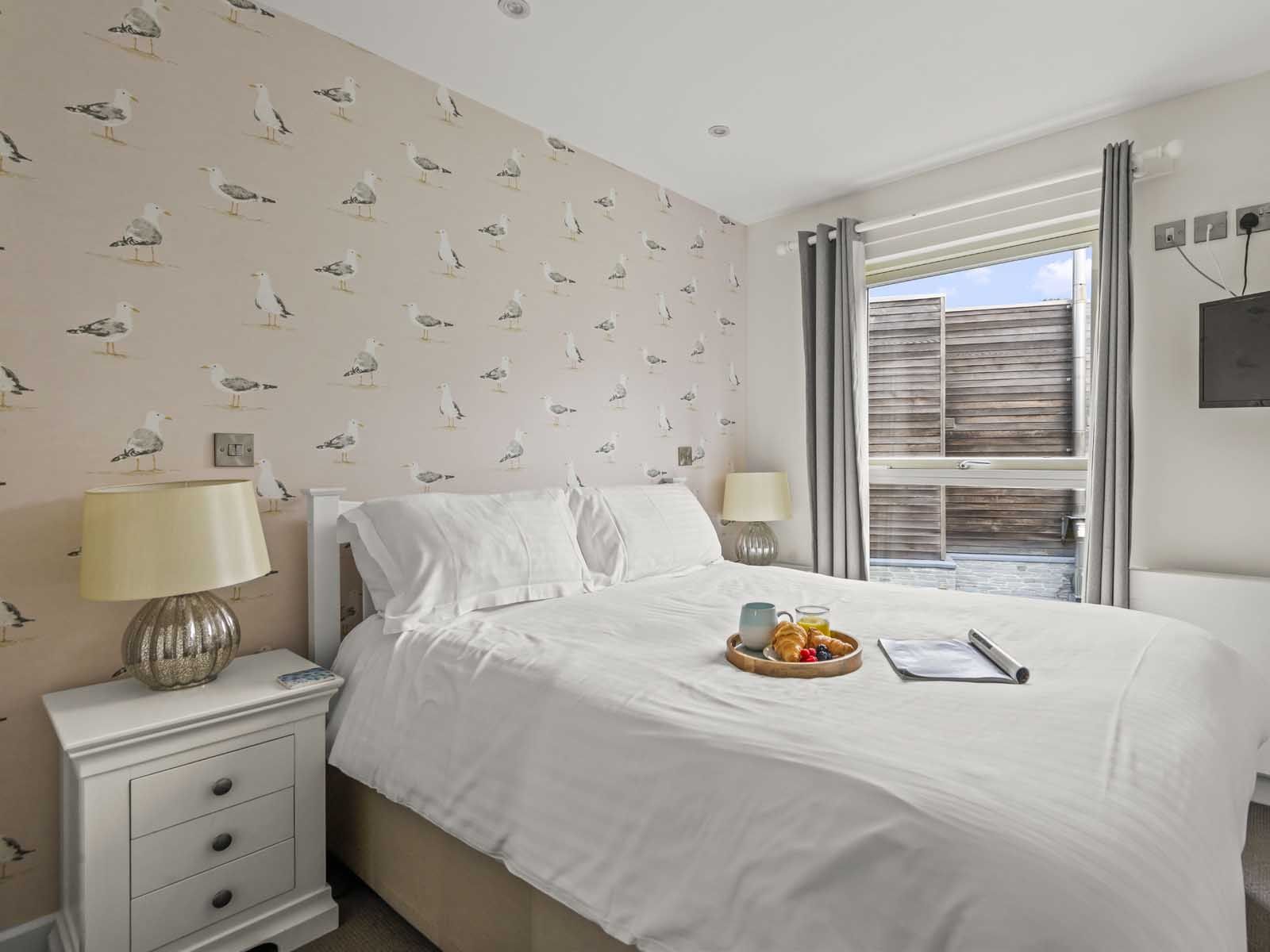 Bright bedroom with seagull wallpaper, white bed linens, tray with breakfast, and mounted TV.
