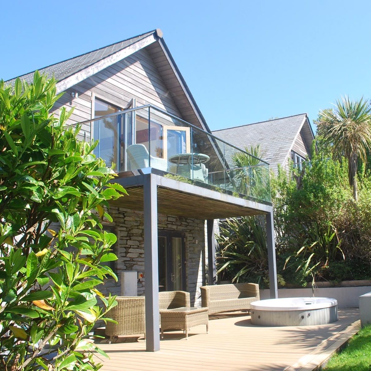 Modern house with glass balcony, patio furniture, and hot tub surrounded by greenery