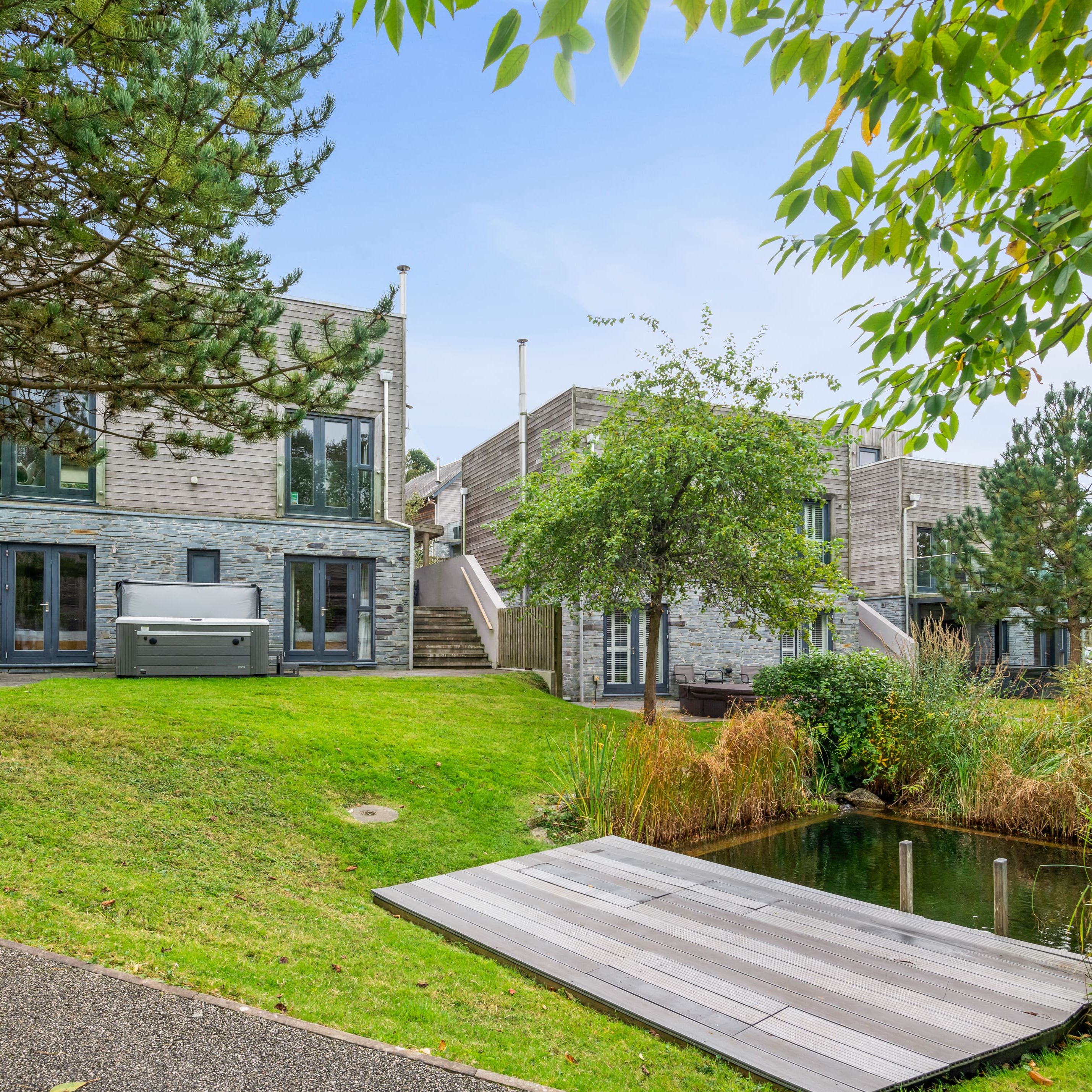Modern houses with stone and wood exteriors, private patios, hot tub, and lush green landscaping with a small pond and dock in the foreground.