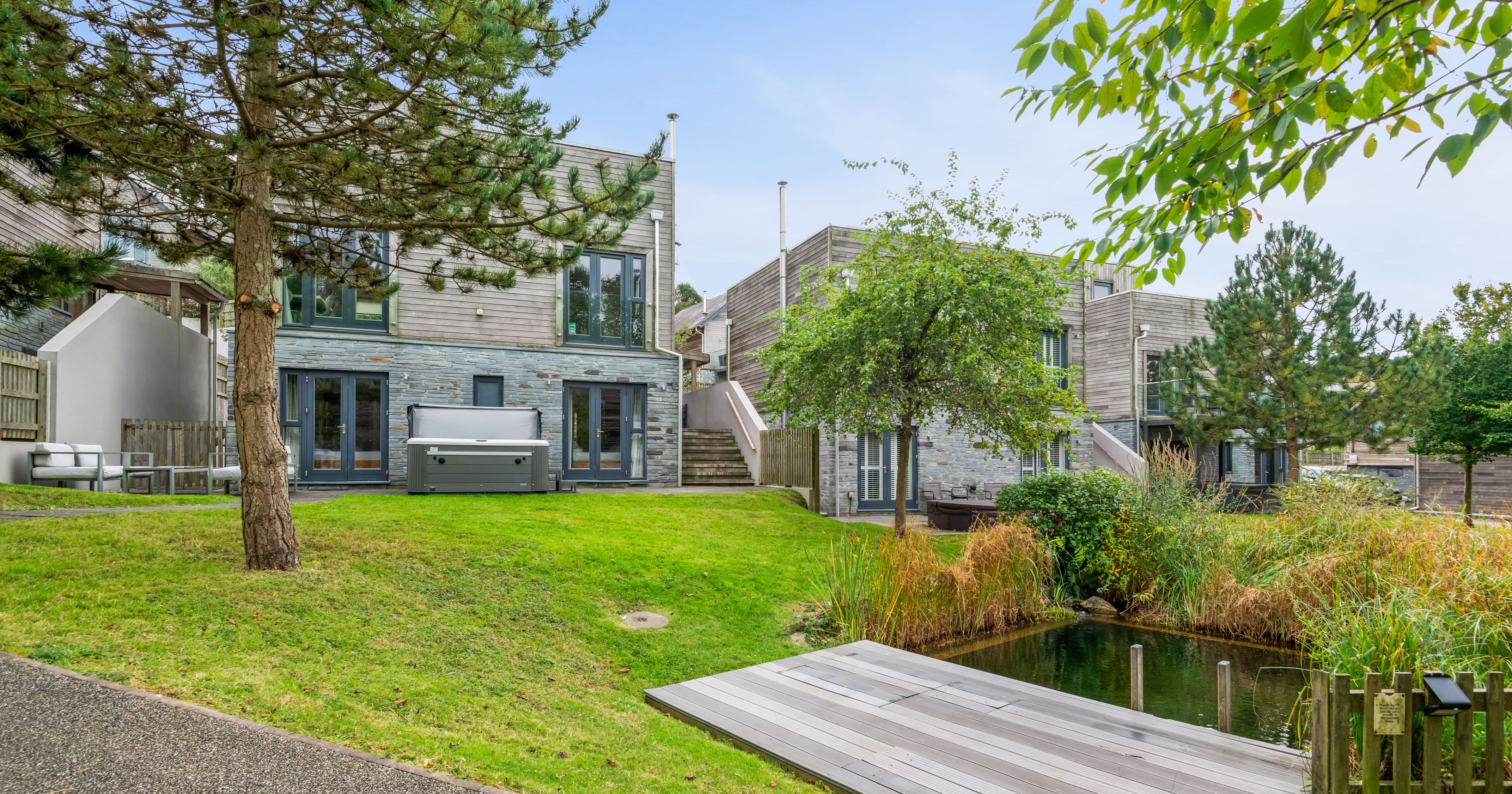 Modern houses with stone and wood exteriors, private patios, hot tub, and lush green landscaping with a small pond and dock in the foreground.