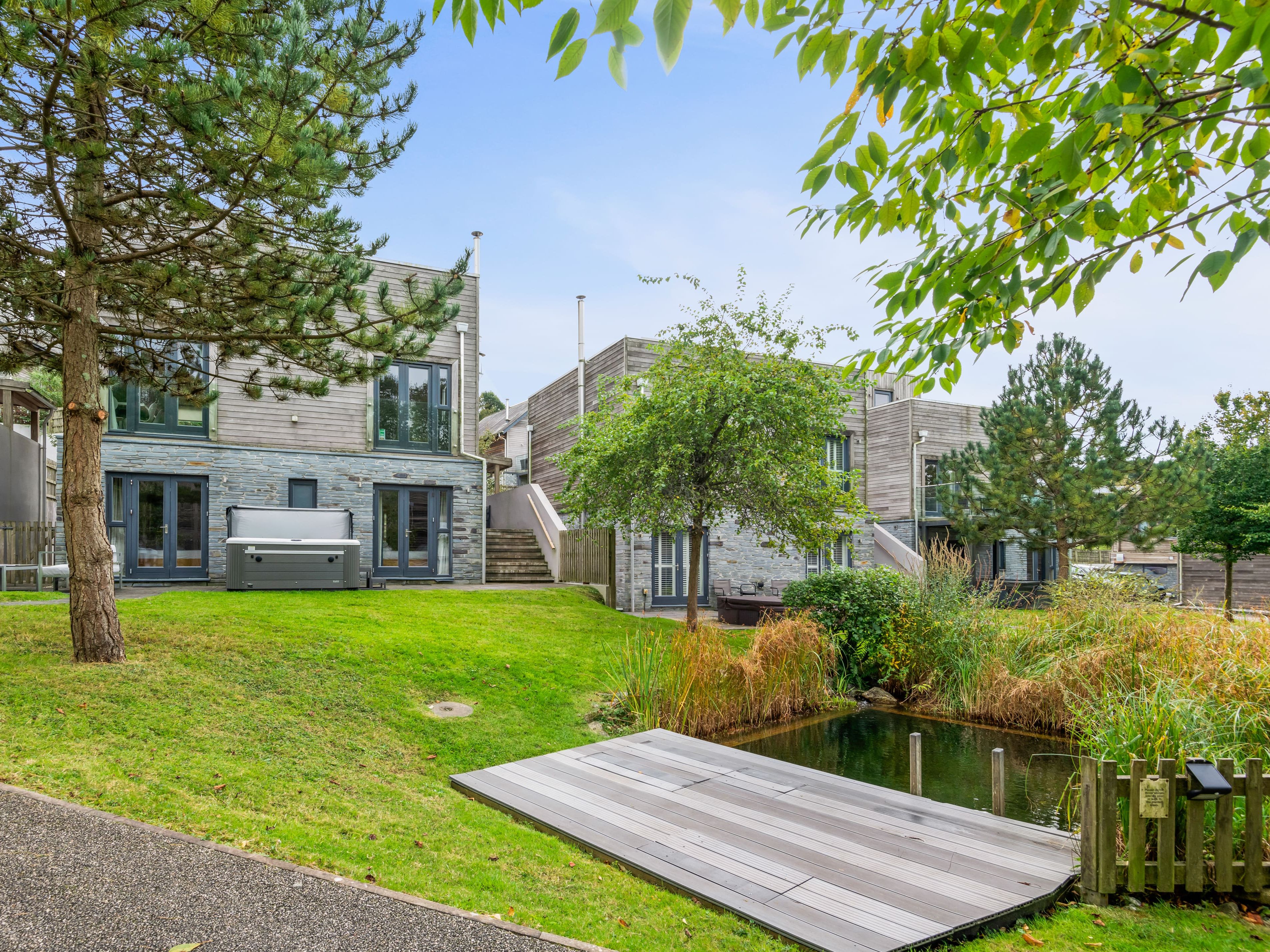 Modern houses with stone and wood exteriors, private patios, hot tub, and lush green landscaping with a small pond and dock in the foreground.