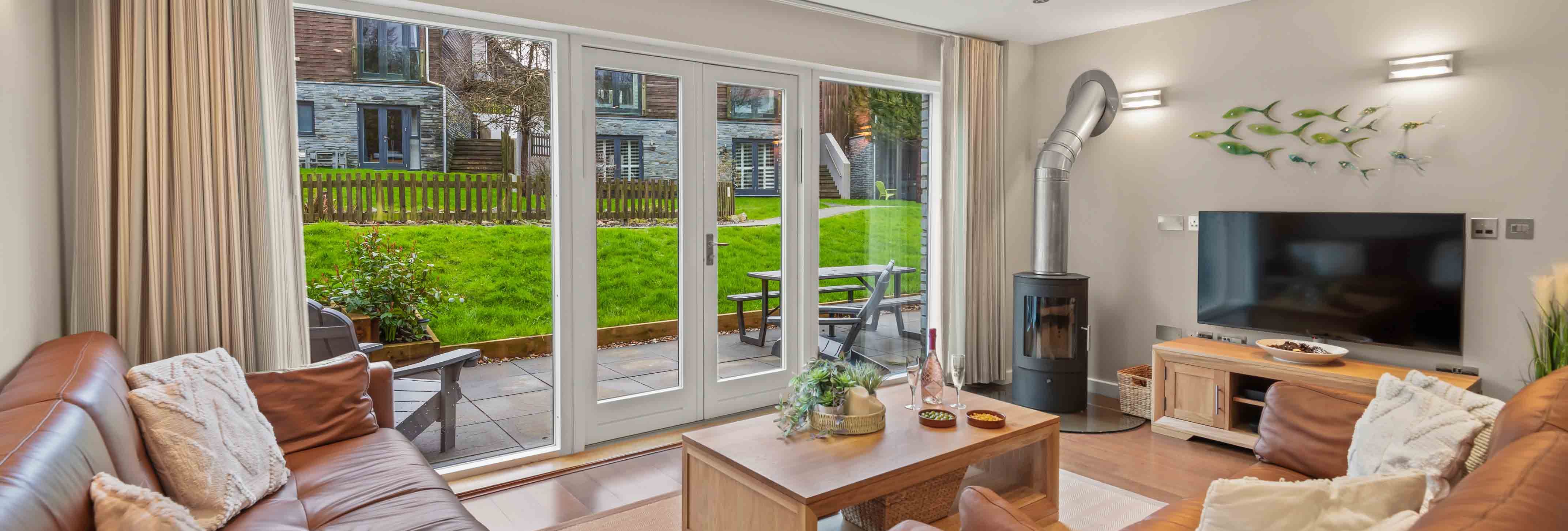 Modern living room with brown leather sofas, wood fireplace, and large glass doors leading to a green outdoor area.