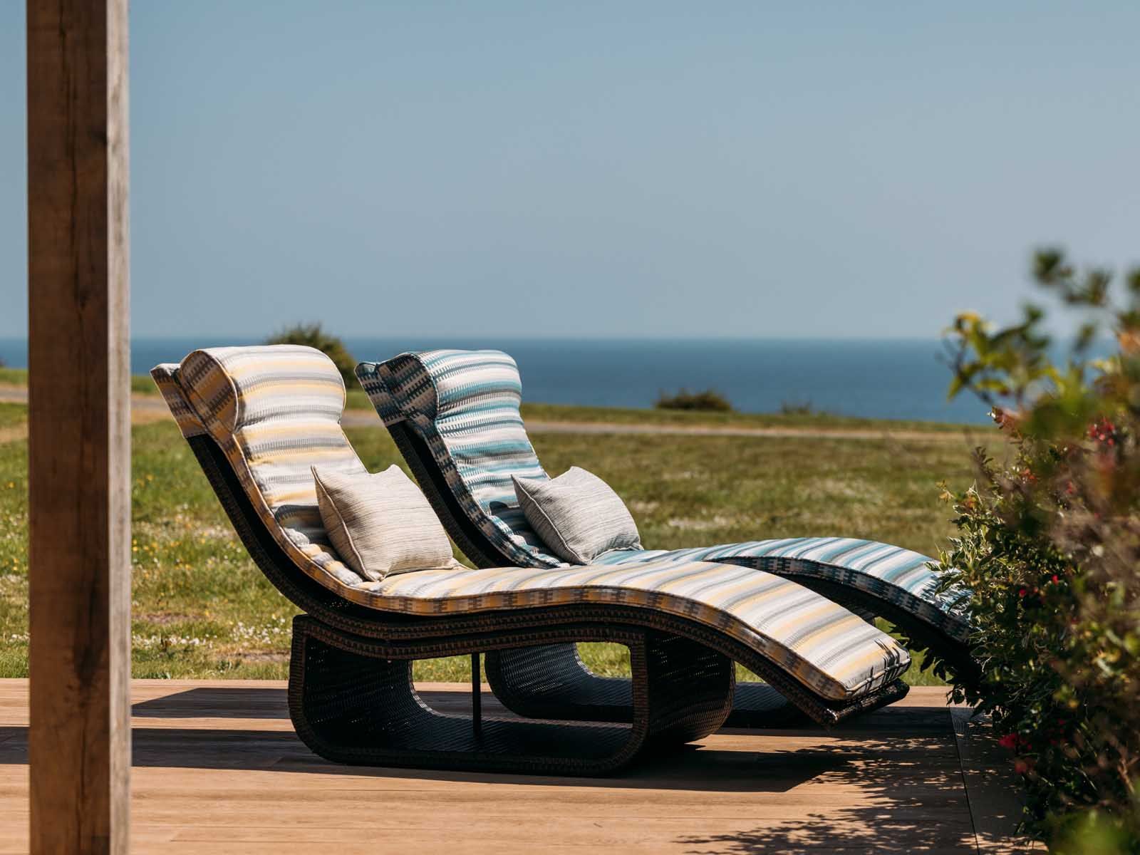 Two striped lounge chairs with cushions on a wooden deck overlooking a grassy area and the sea.