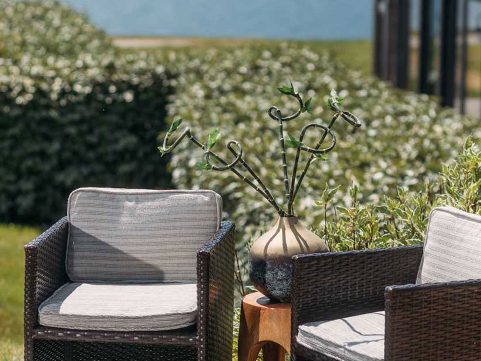 Outdoor wicker chairs with cushions and a decorative vase on a small wooden table, set in a garden with lush greenery.