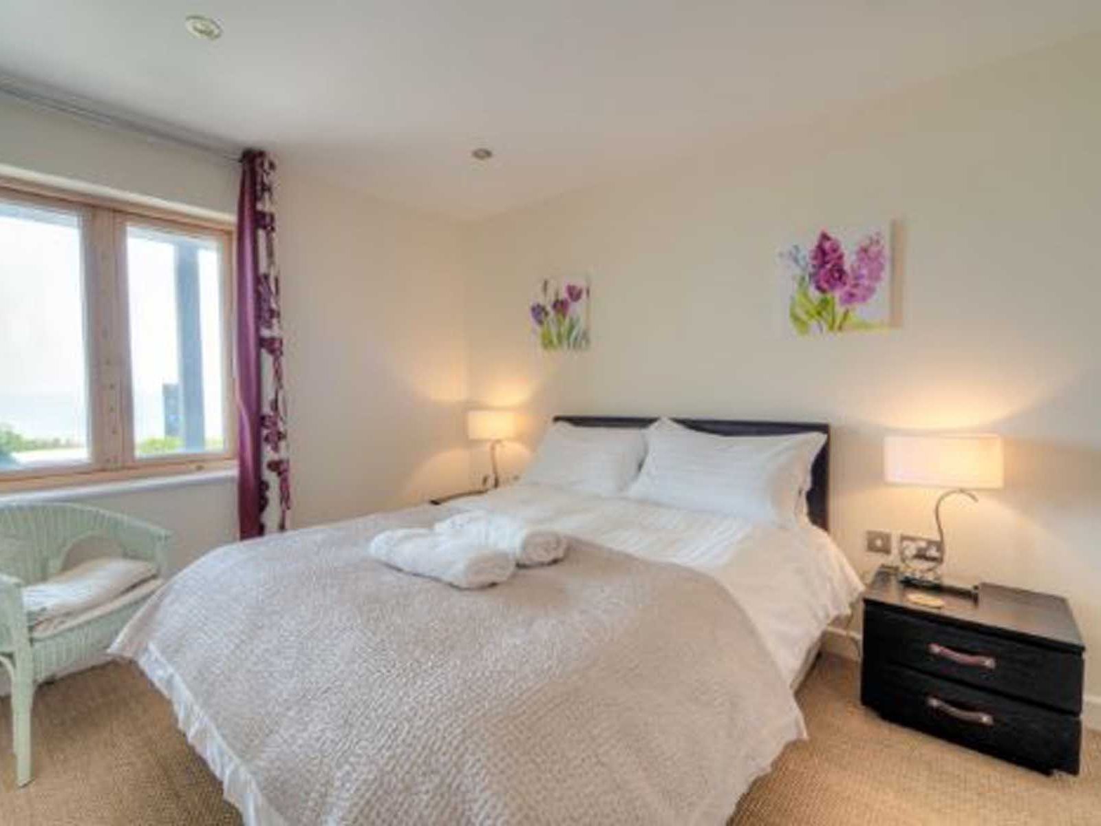 Bright bedroom with double bed, bedside tables, lamps, wicker chair, and window with purple curtains