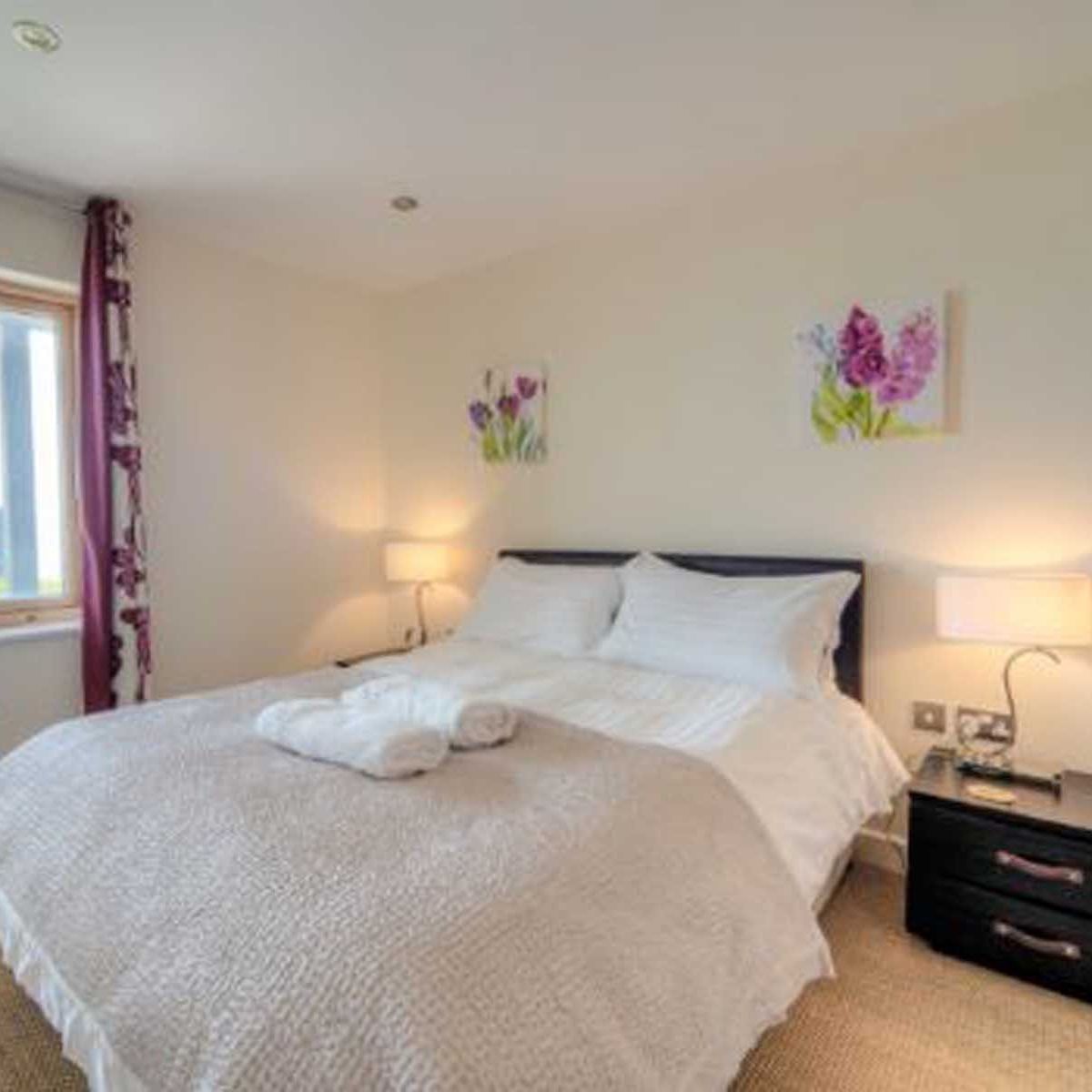 Bright bedroom with double bed, bedside tables, lamps, wicker chair, and window with purple curtains