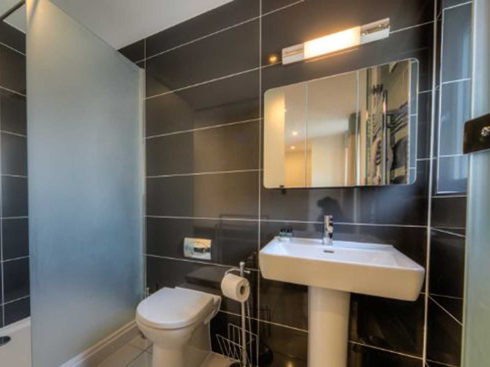 Modern bathroom with black tiled walls, pedestal sink, toilet, and frosted glass shower partition