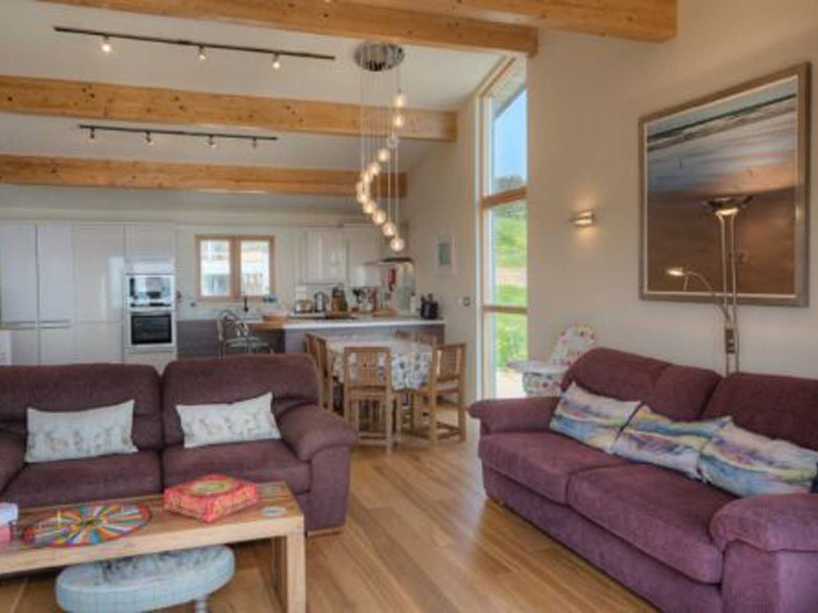 Modern open-plan living room and kitchen with wooden beams, purple sofas, dining table, and a large window.