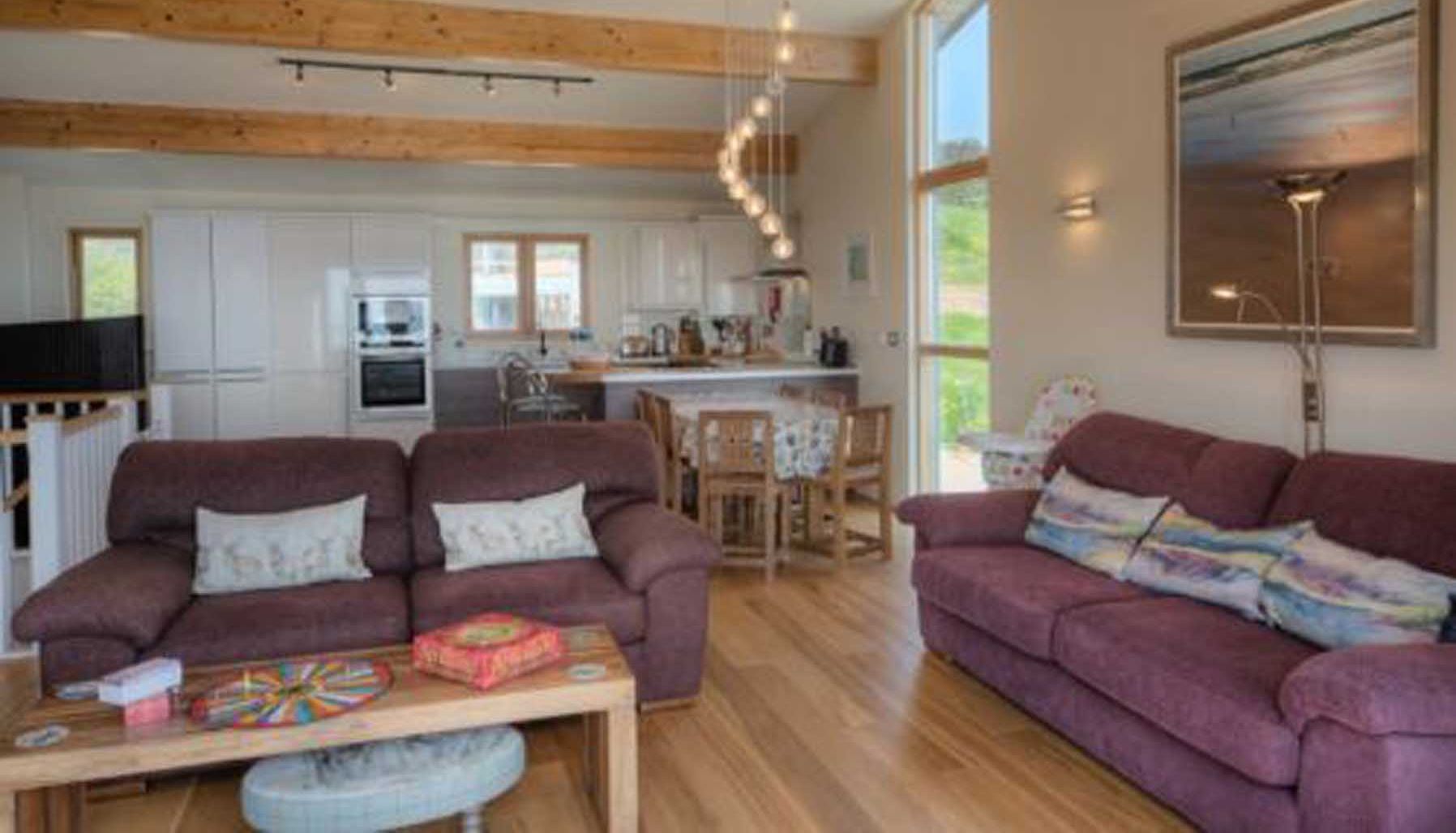 Modern open-plan living room and kitchen with wooden beams, purple sofas, dining table, and a large window.