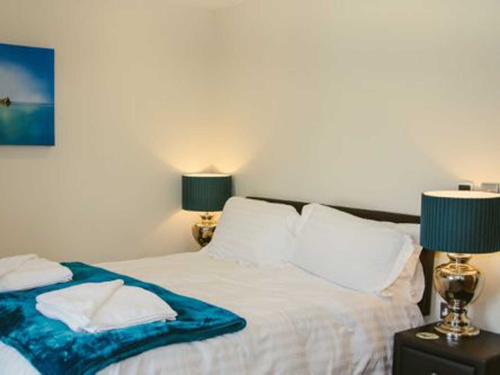 Modern bedroom with white bedding, teal blanket and lamps, and a seascape painting on the wall