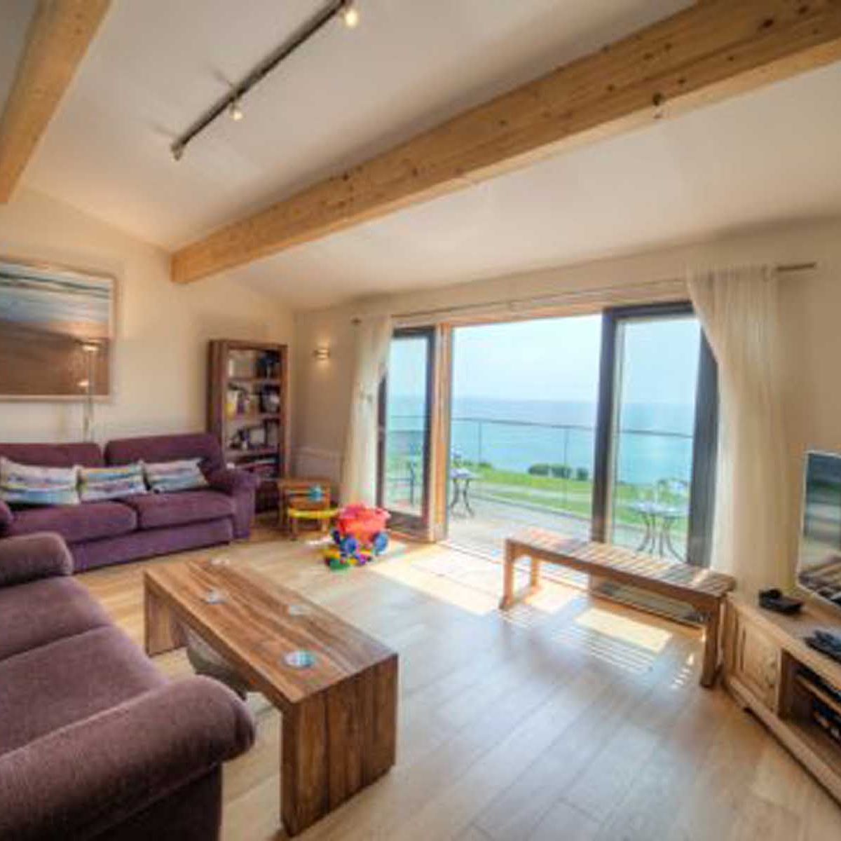 Bright living room with large windows overlooking the ocean, featuring purple sofas, wooden furniture, and children's toys.