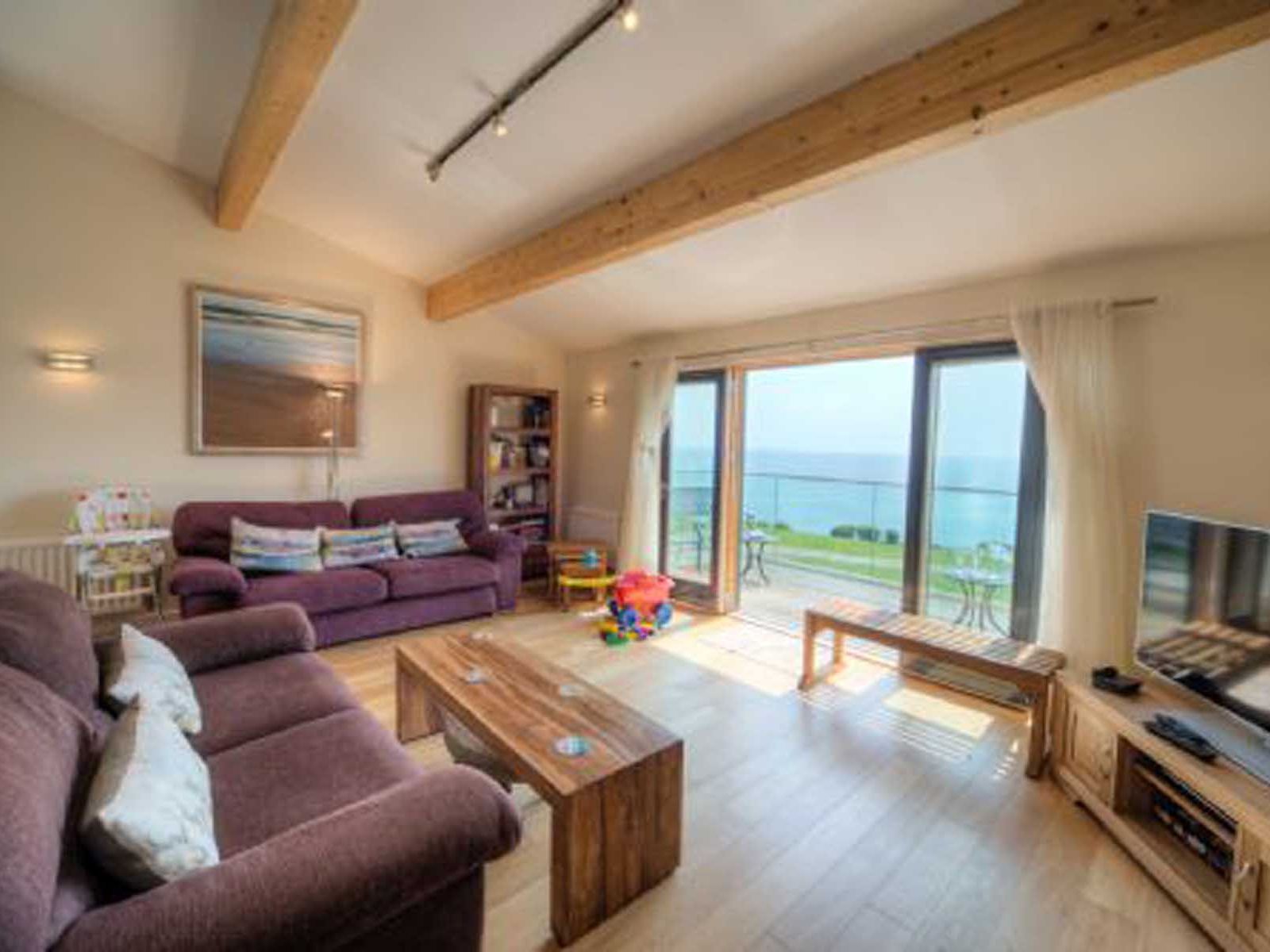 Bright living room with large windows overlooking the ocean, featuring purple sofas, wooden furniture, and children's toys.