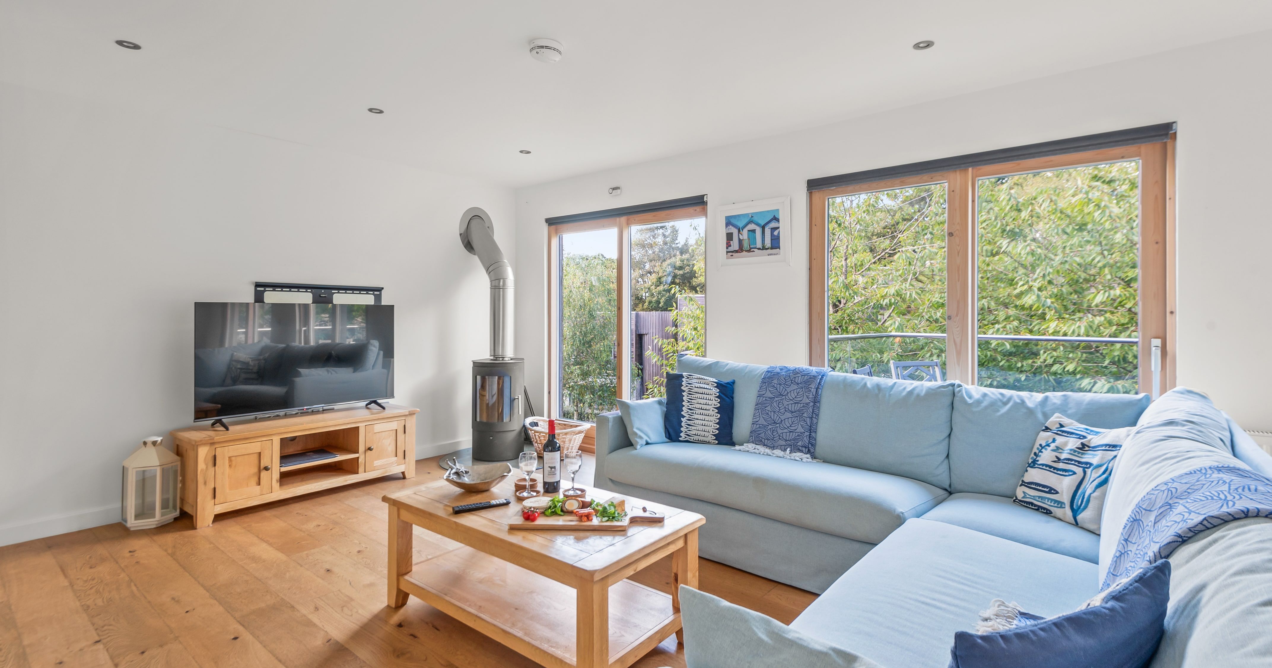 Bright and modern living room with large windows, light blue sectional sofa, wooden coffee table, television on a wooden stand, and a wood-burning stove.