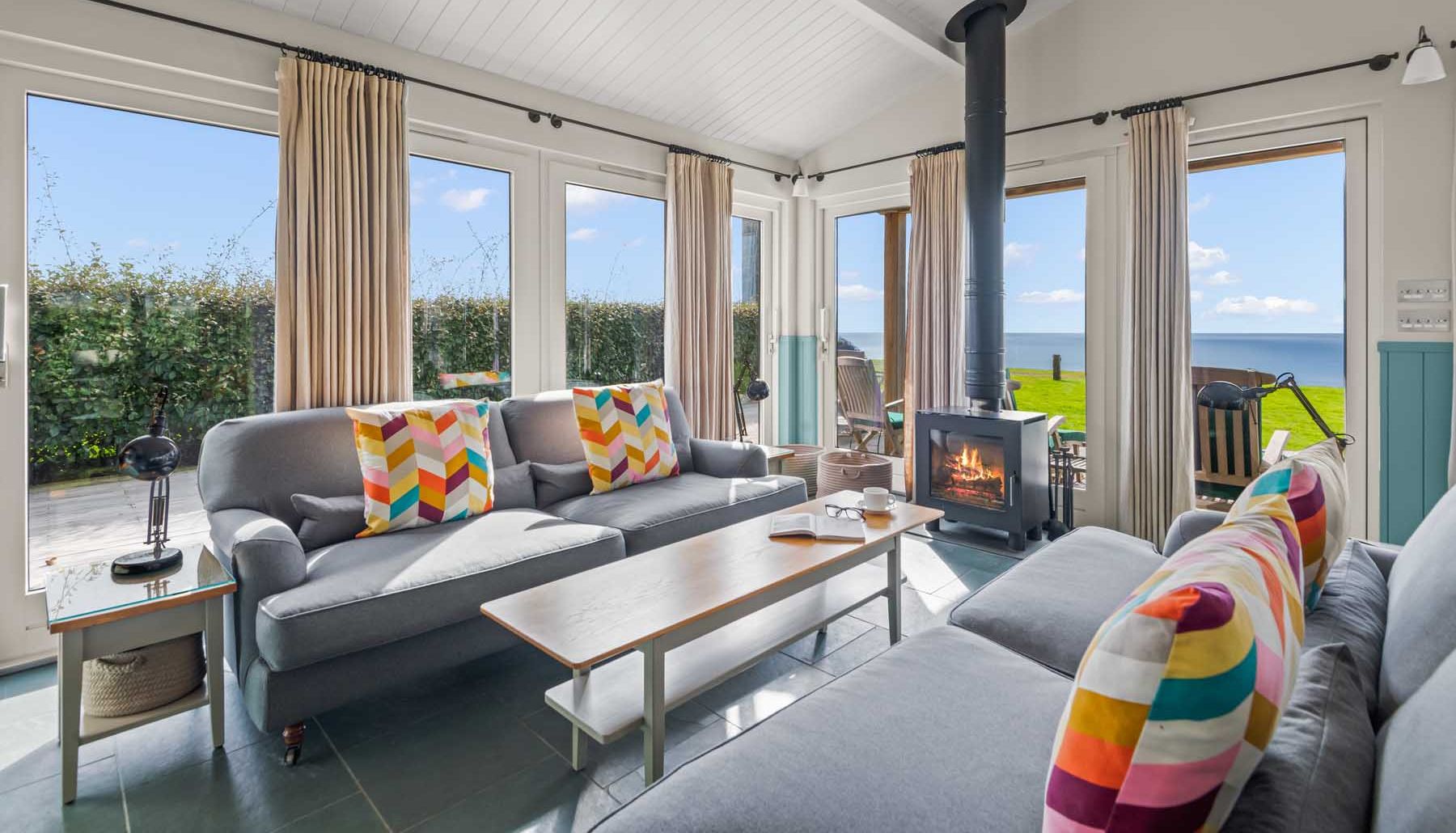 Bright and cozy living room with colorful pillows, wood-burning stove, and ocean view through large windows.