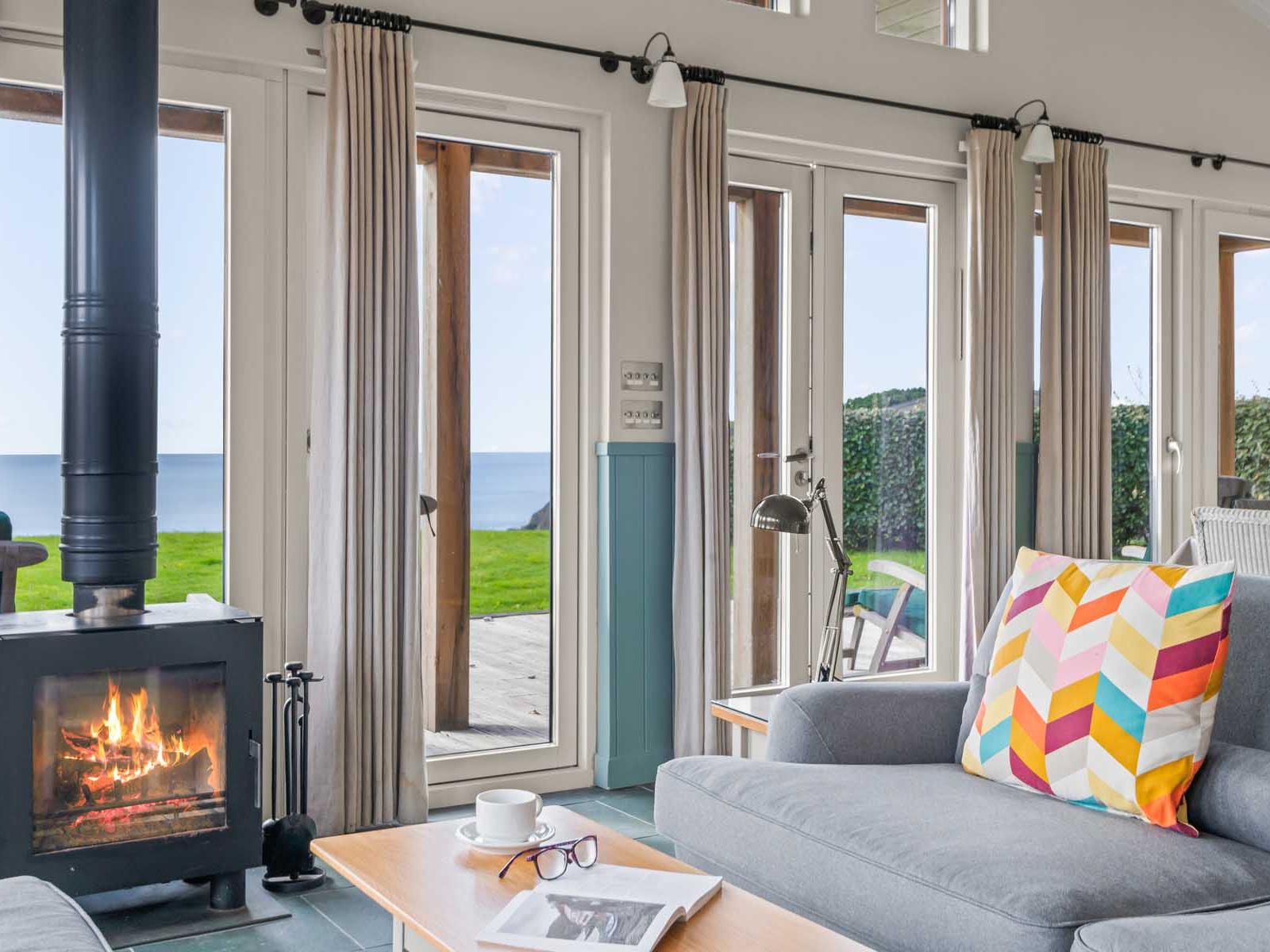 Cozy living room with wood burning stove, colorful cushion, and large windows overlooking the ocean.