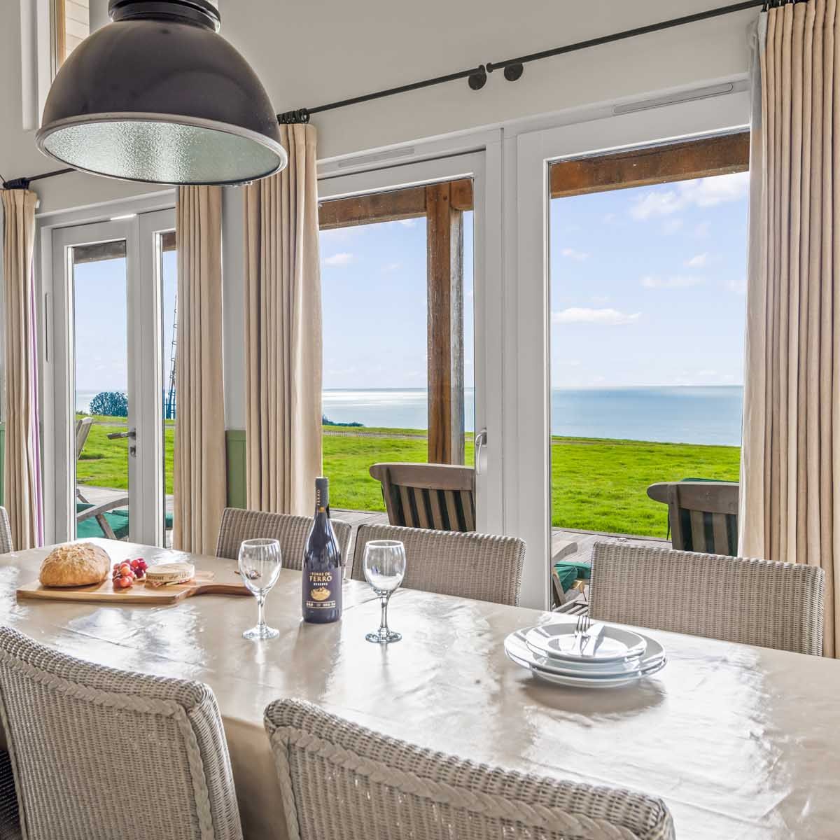 Dining table in a bright room with large windows overlooking a grassy field and the sea.