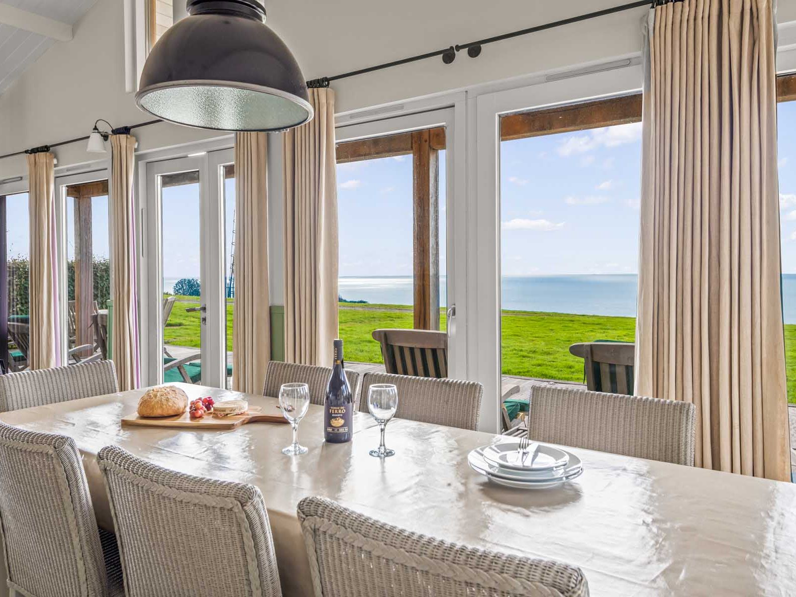 Dining table in a bright room with large windows overlooking a grassy field and the sea.