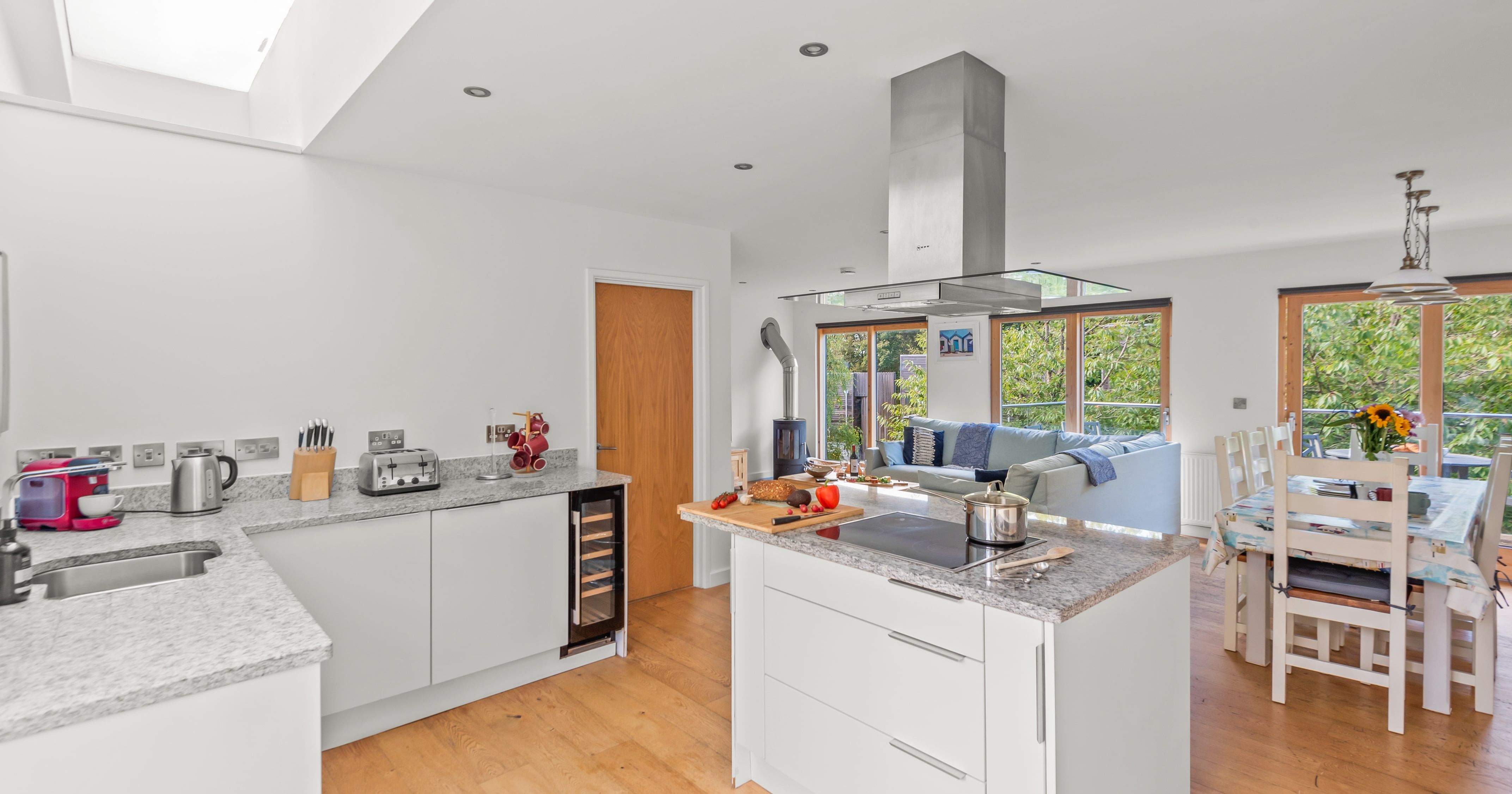 Bright, modern open-plan kitchen and living area with white cabinets, granite countertops, central island with stove, wooden floors, dining table, and large windows overlooking greenery.