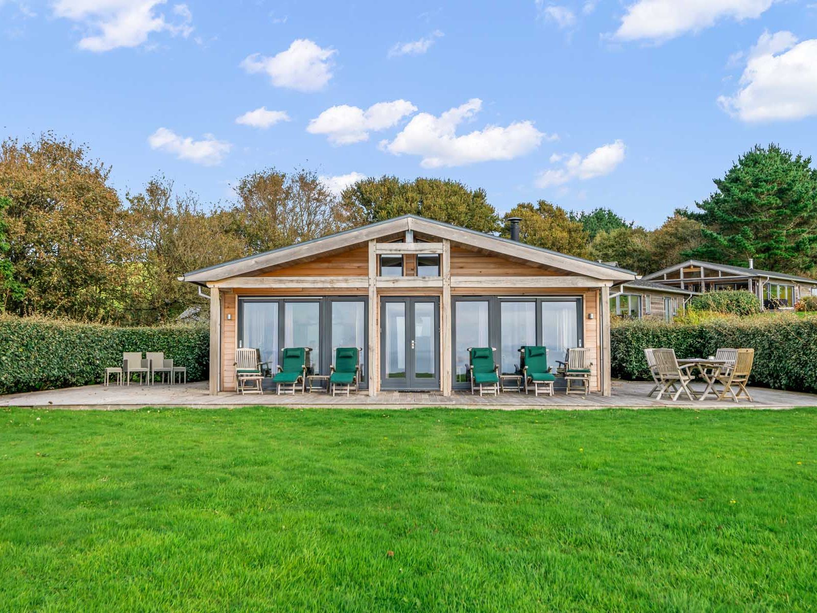 Modern wooden cabin with patio chairs, tables, and a green lawn in front.