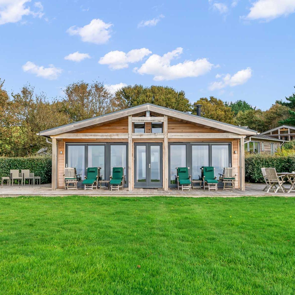 Modern wooden cabin with patio chairs, tables, and a green lawn in front.