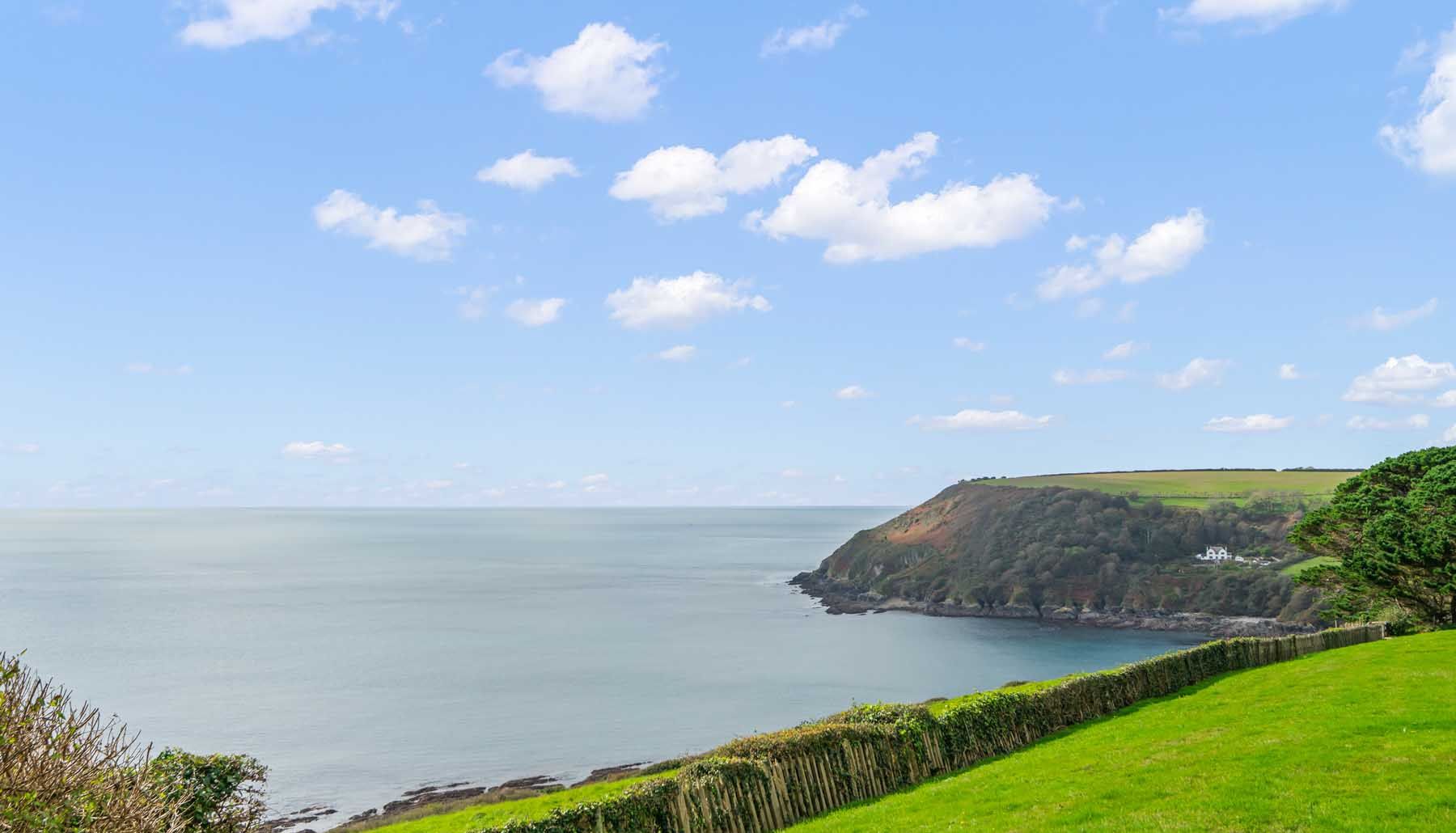 Coastal landscape with grassy hill, wooden fence, and ocean under a blue sky with scattered clouds.
