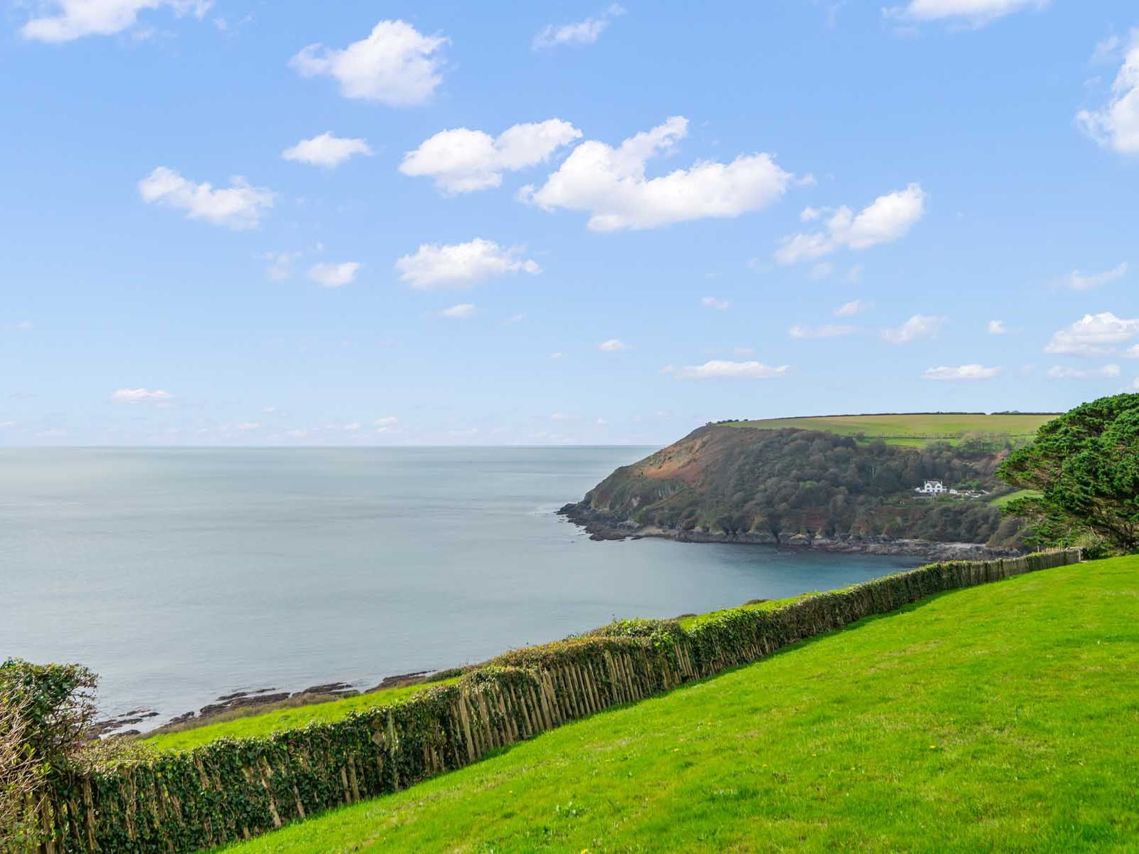 Coastal landscape with grassy hill, wooden fence, and ocean under a blue sky with scattered clouds.