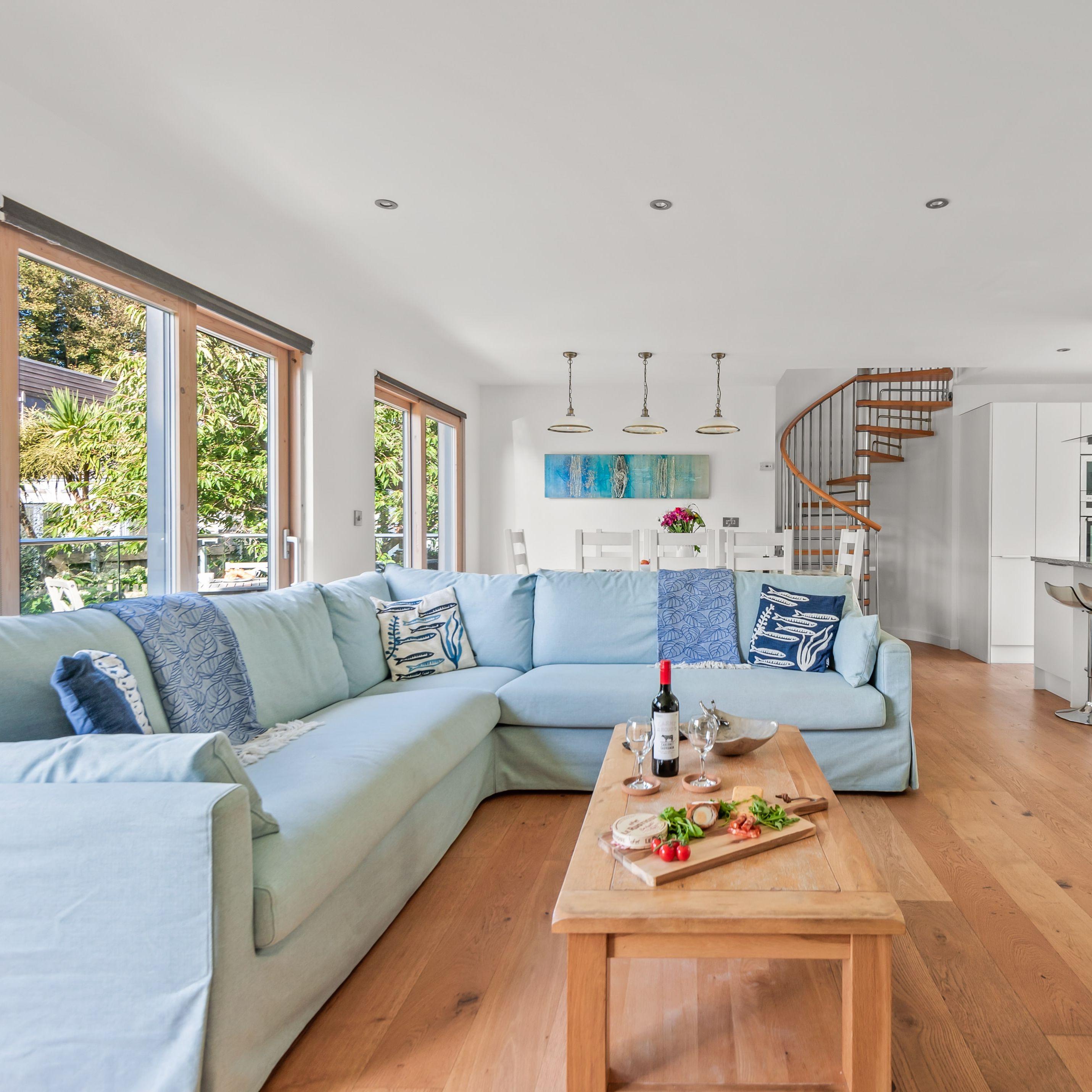 Modern open-plan living room and kitchen with light blue sectional sofa, wooden floor, large windows, and spiral staircase.