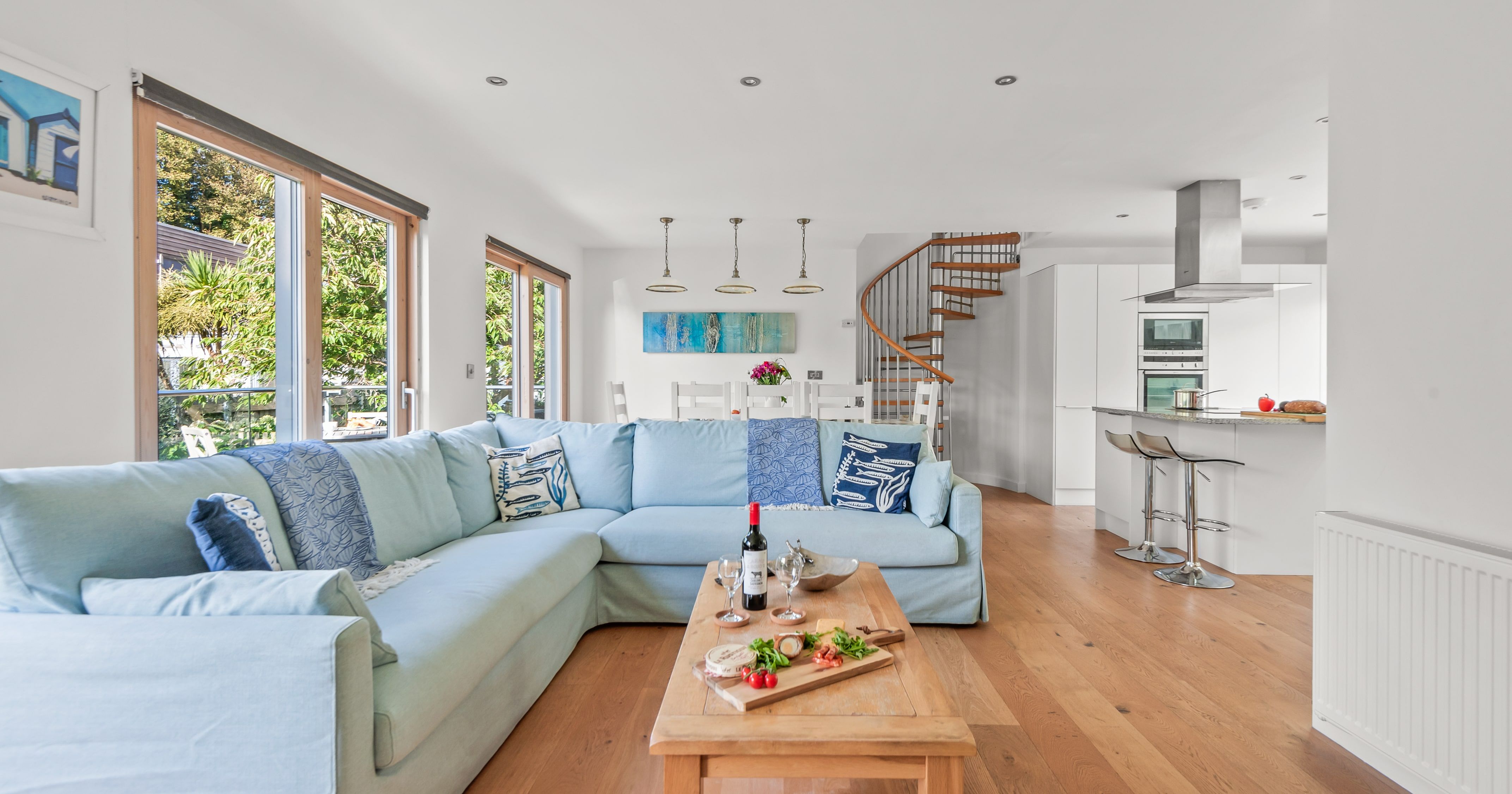 Modern open-plan living room and kitchen with light blue sectional sofa, wooden floor, large windows, and spiral staircase.