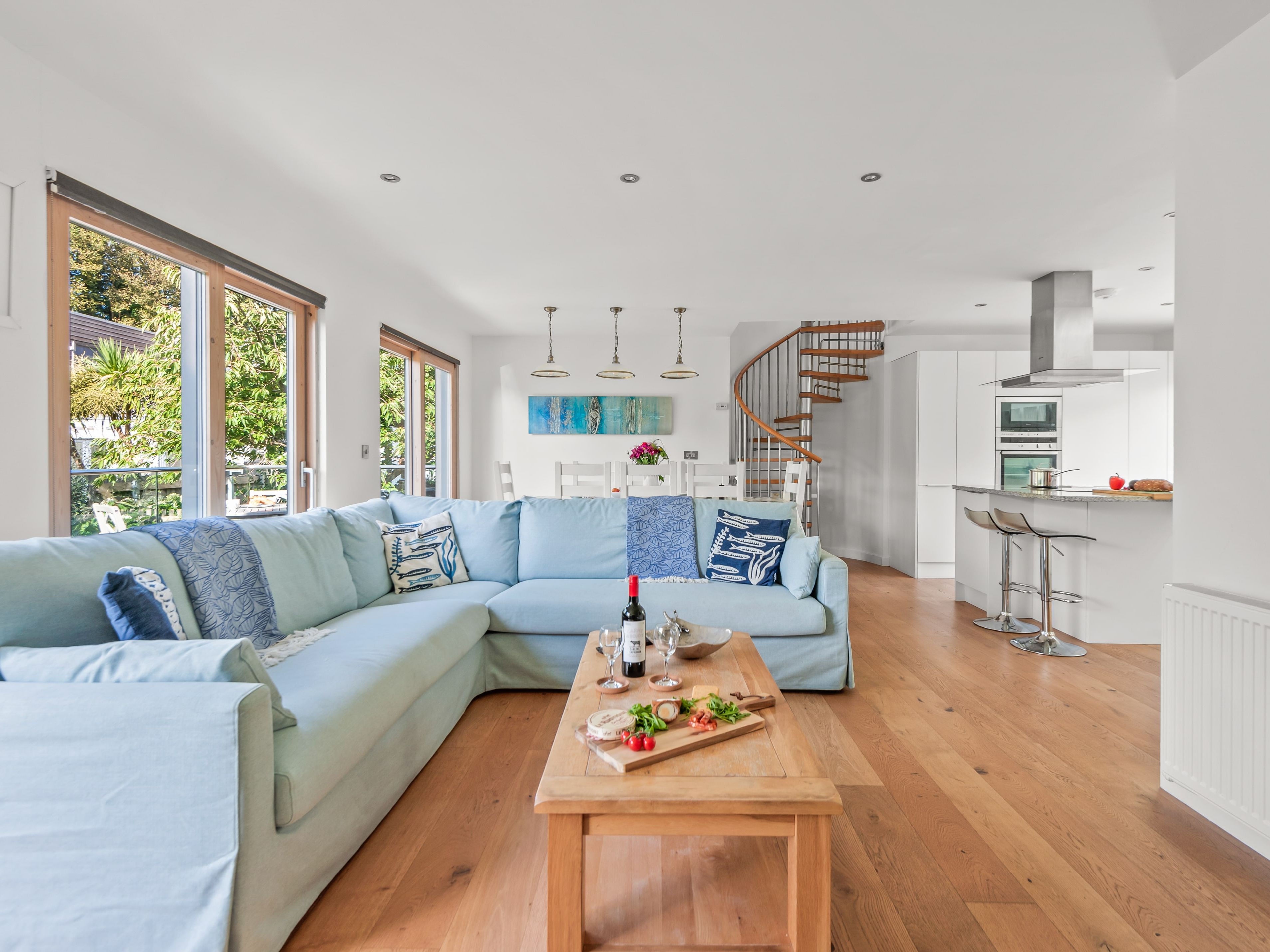 Modern open-plan living room and kitchen with light blue sectional sofa, wooden floor, large windows, and spiral staircase.