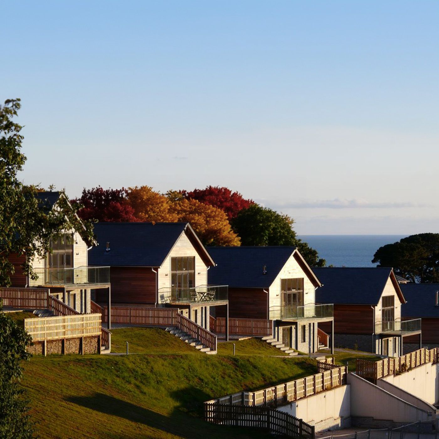 Modern wooden houses with glass balconies on a hillside overlooking the sea, surrounded by autumn trees.
