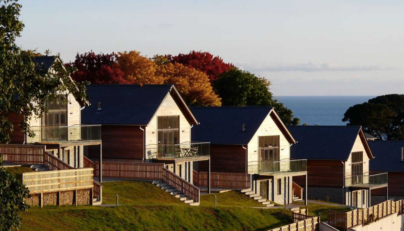 Modern wooden houses with glass balconies on a hillside overlooking the sea, surrounded by autumn trees.