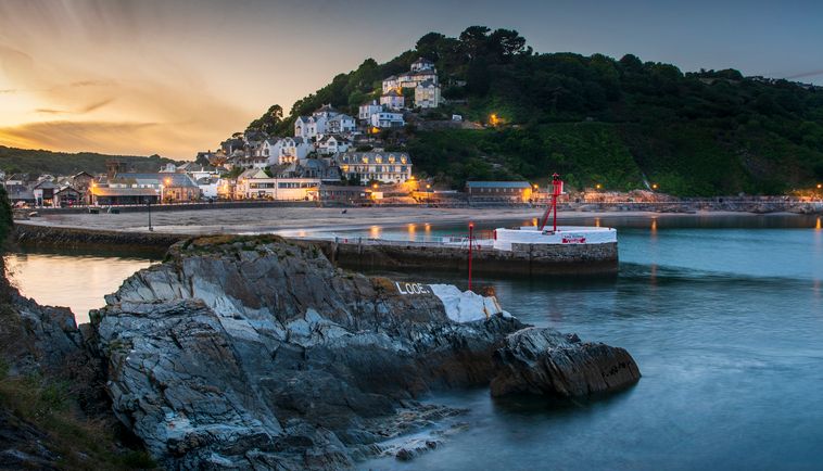 Coastal village with illuminated houses on a hillside overlooking a calm harbor at dusk.