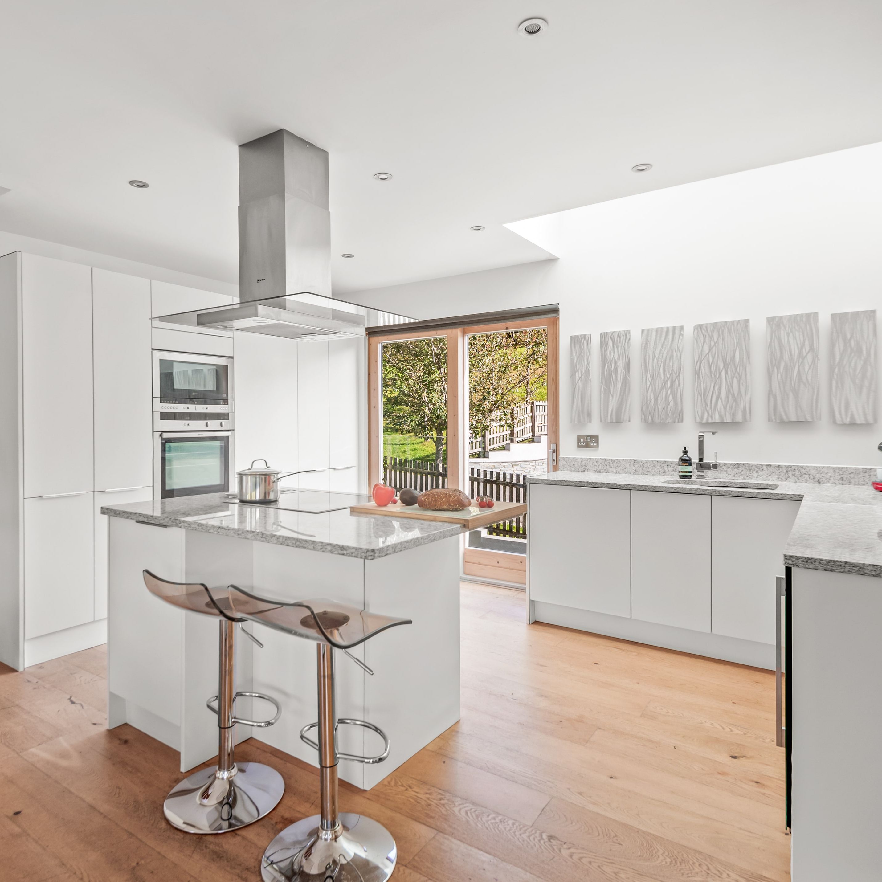 Modern white kitchen with granite countertops, wooden floors, a spiral staircase, kitchen island with stools, large windows, and contemporary decor.