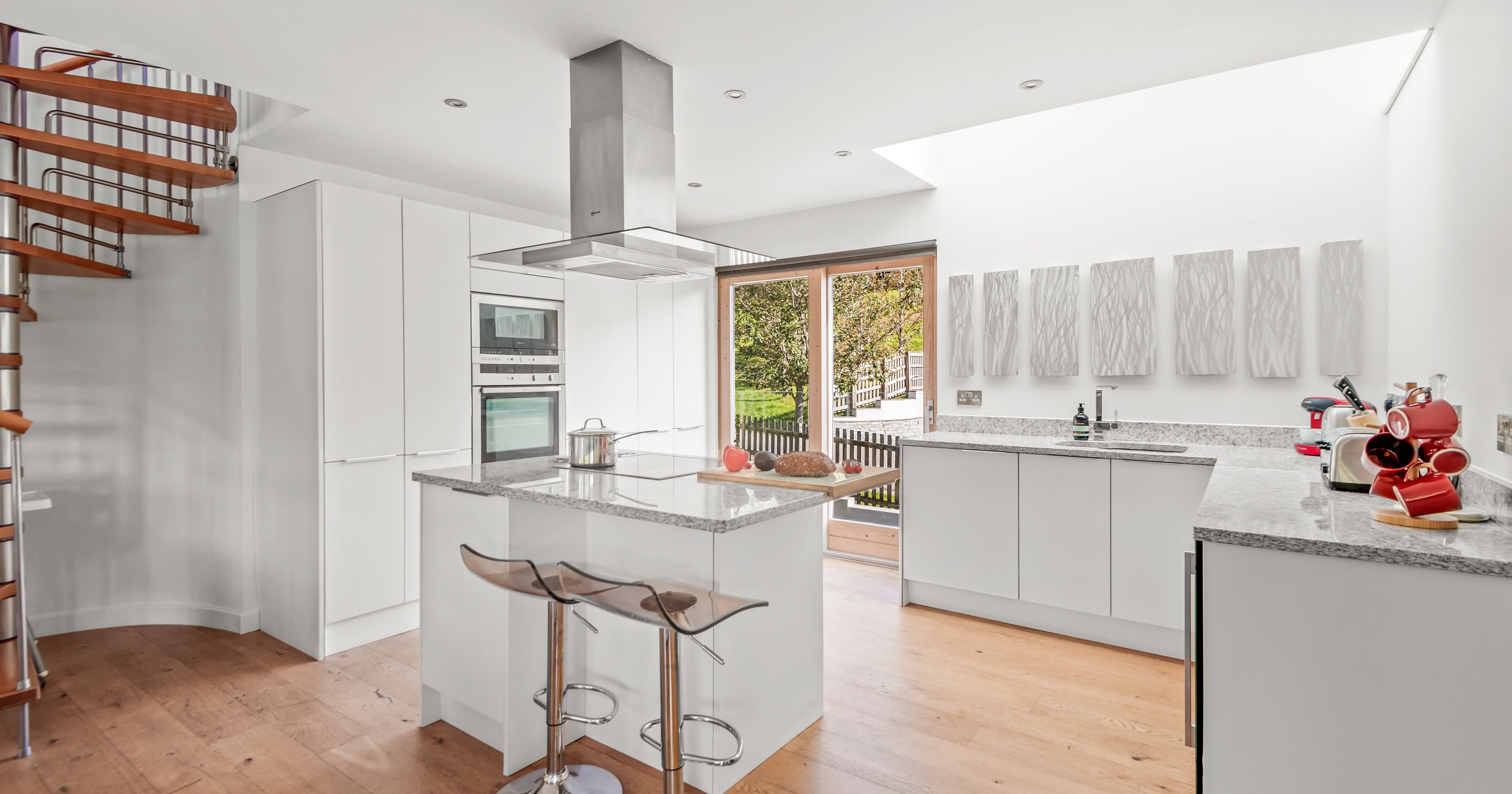 Modern white kitchen with granite countertops, wooden floors, a spiral staircase, kitchen island with stools, large windows, and contemporary decor.
