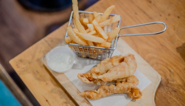 Basket of French fries with battered fish fillets and dipping sauce on a wooden board.