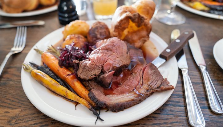 Plate of roast beef with Yorkshire pudding, carrots, potatoes, and red cabbage on a wooden table.