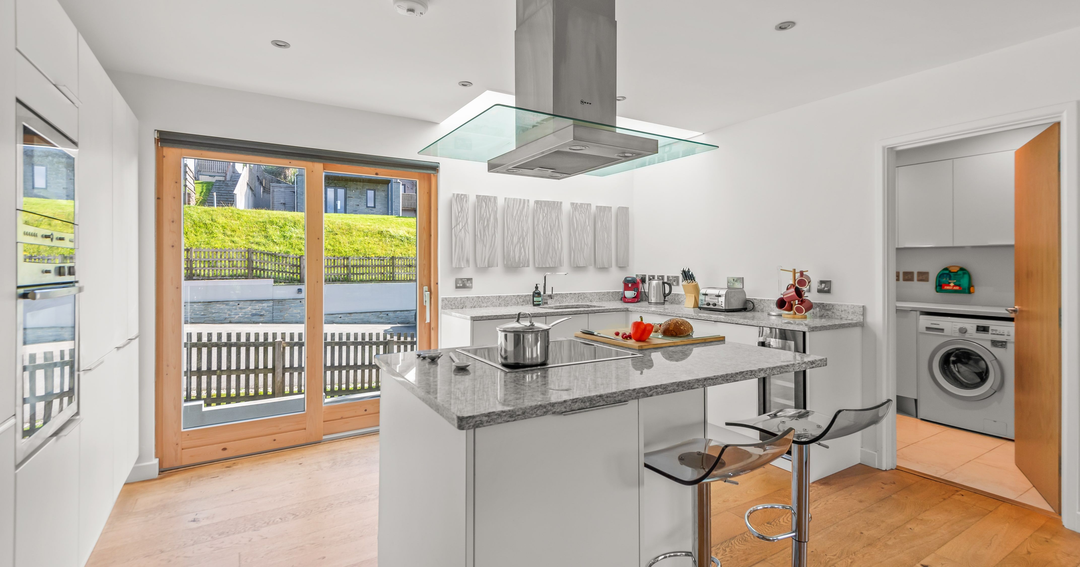 Modern kitchen with sleek white cabinets, granite countertops, a kitchen island with bar stools, and large glass doors letting in natural light. The adjacent room contains a washing machine.