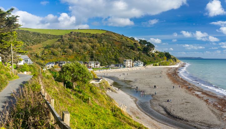Coastal path overlooking a pebbly beach with people, bordered by hillside greenery and houses, under a partly cloudy sky.
