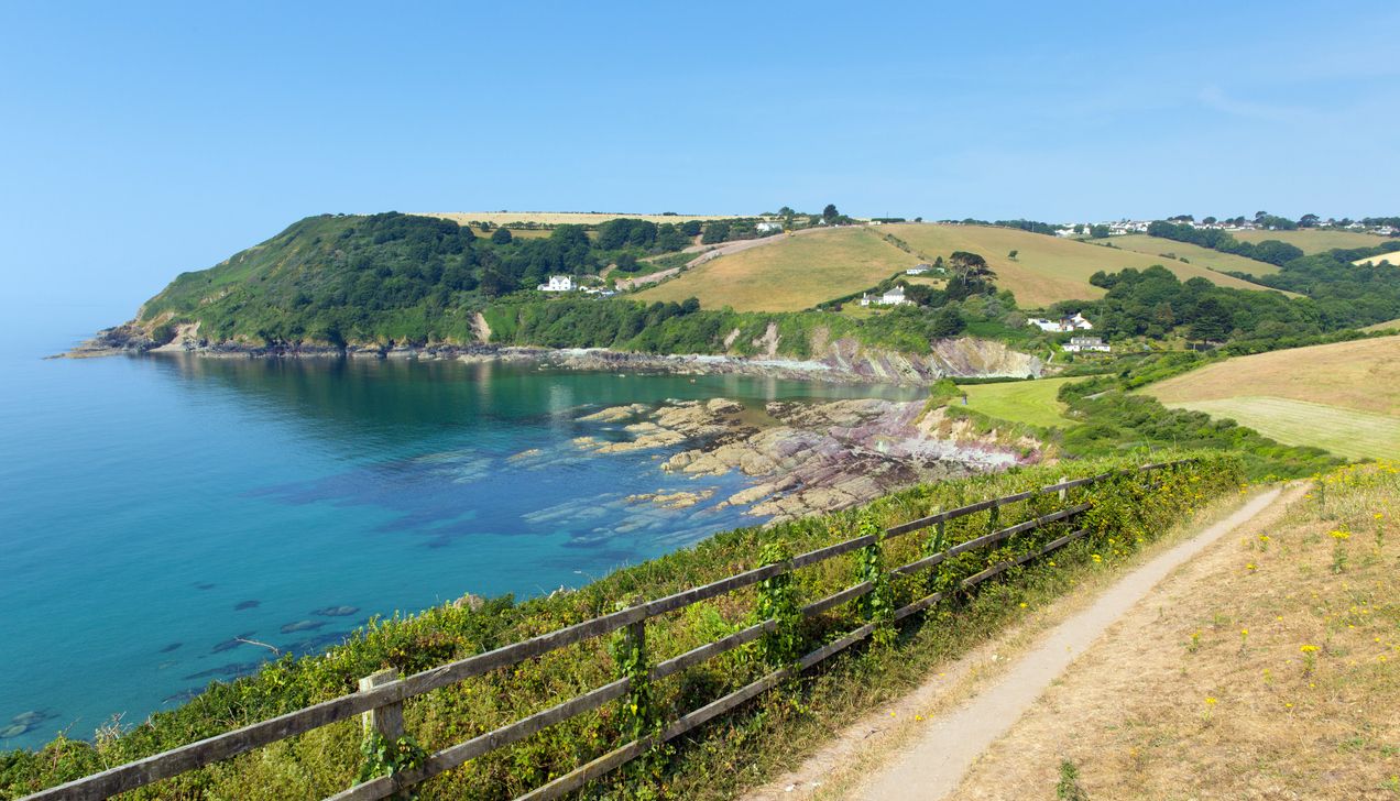 Coastal footpath overlooking a scenic bay with clear blue water, green hills, and countryside homes.