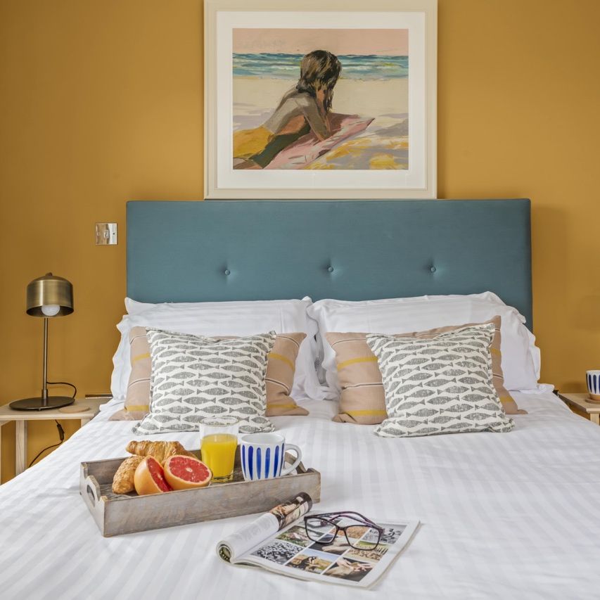 Cozy bedroom with mustard yellow walls, a teal upholstered headboard, two bedside tables with lamps, and a breakfast tray on a neatly made bed.