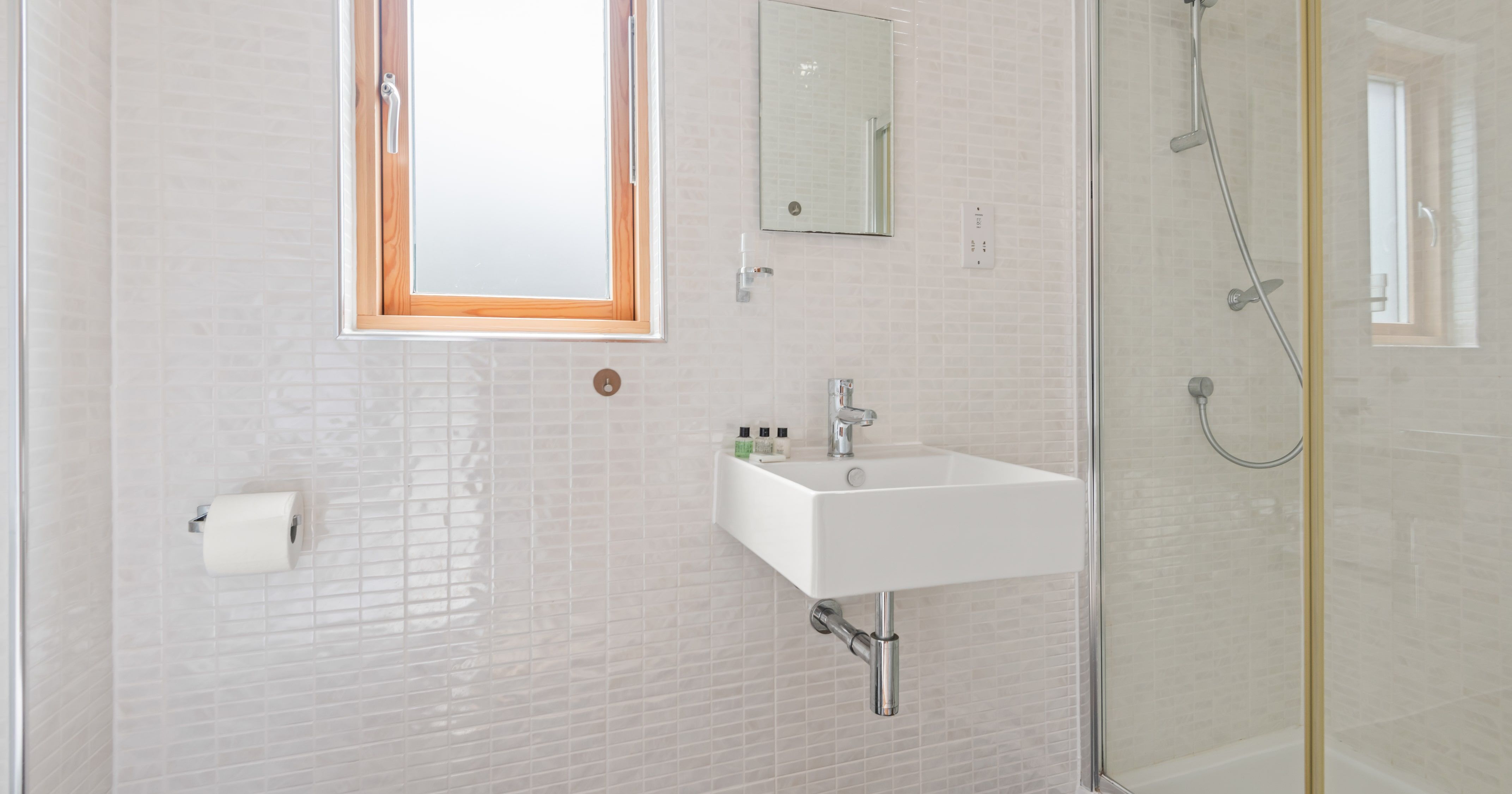 Modern white bathroom with a small square sink, glass-enclosed shower, frosted window, and wall-mounted mirror.