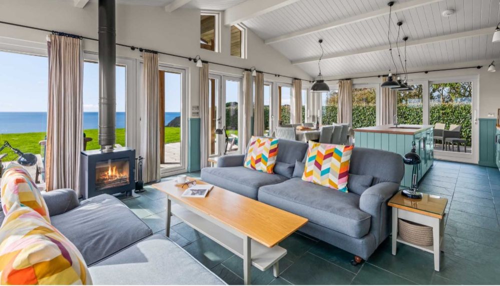 Modern living room with large windows overlooking the ocean, featuring colorful pillows, a wood stove, and open kitchen area.